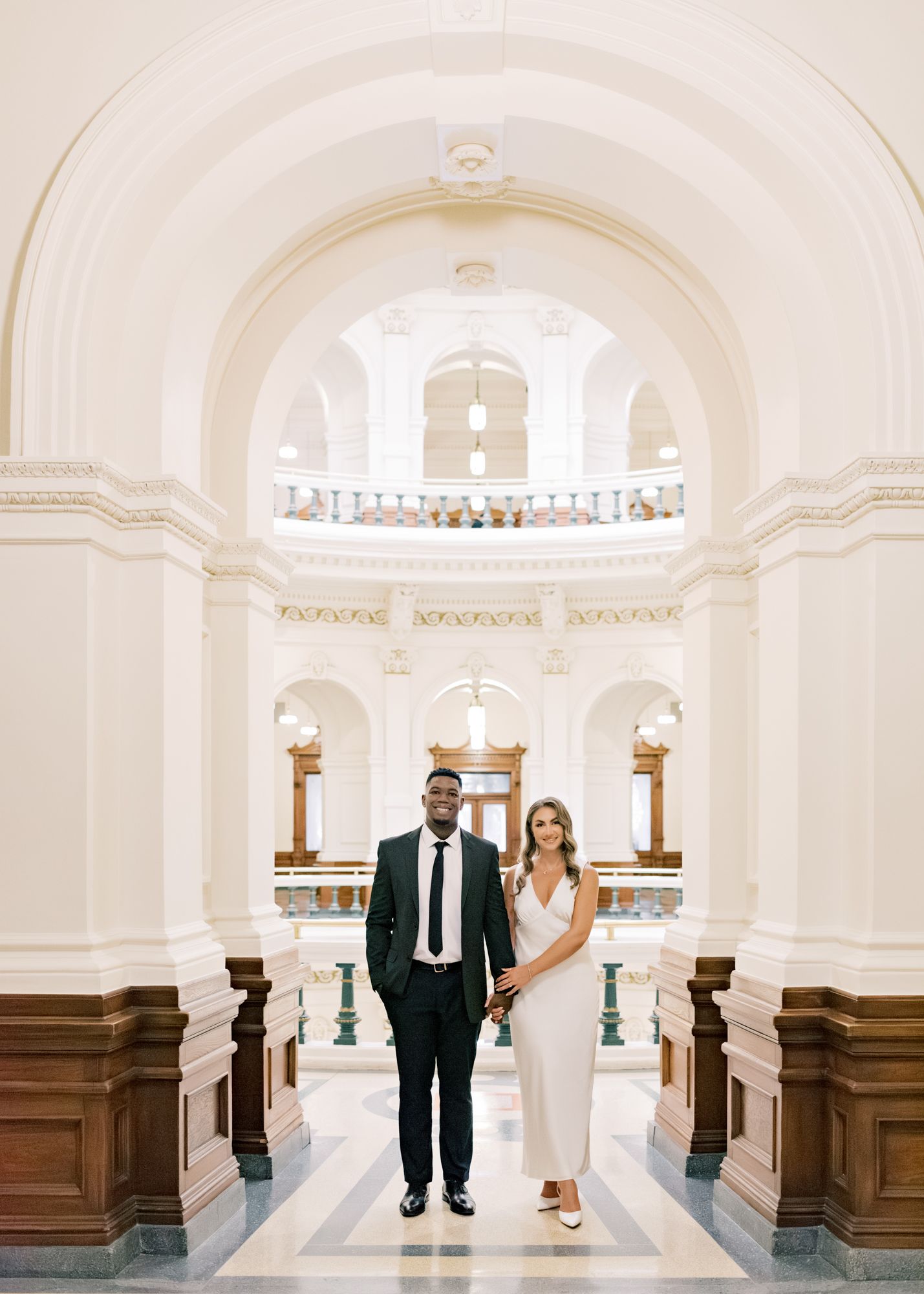 A couple stands in a grand white hallway with arched ceilings and wooden details.