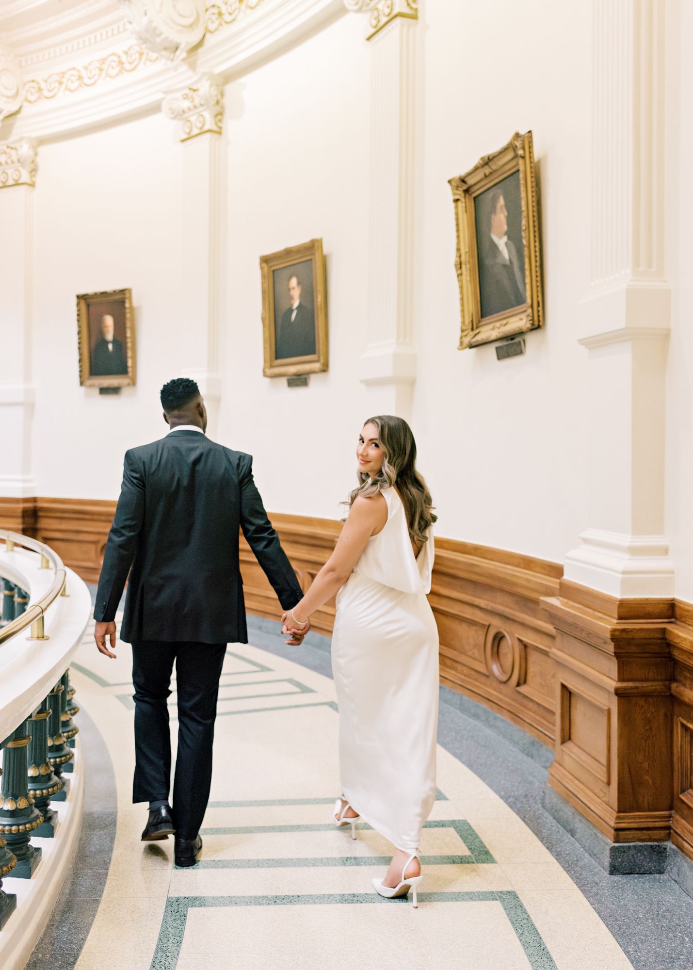 A couple walks hand in hand down an elegant hallway with historic portraits on the wall.
