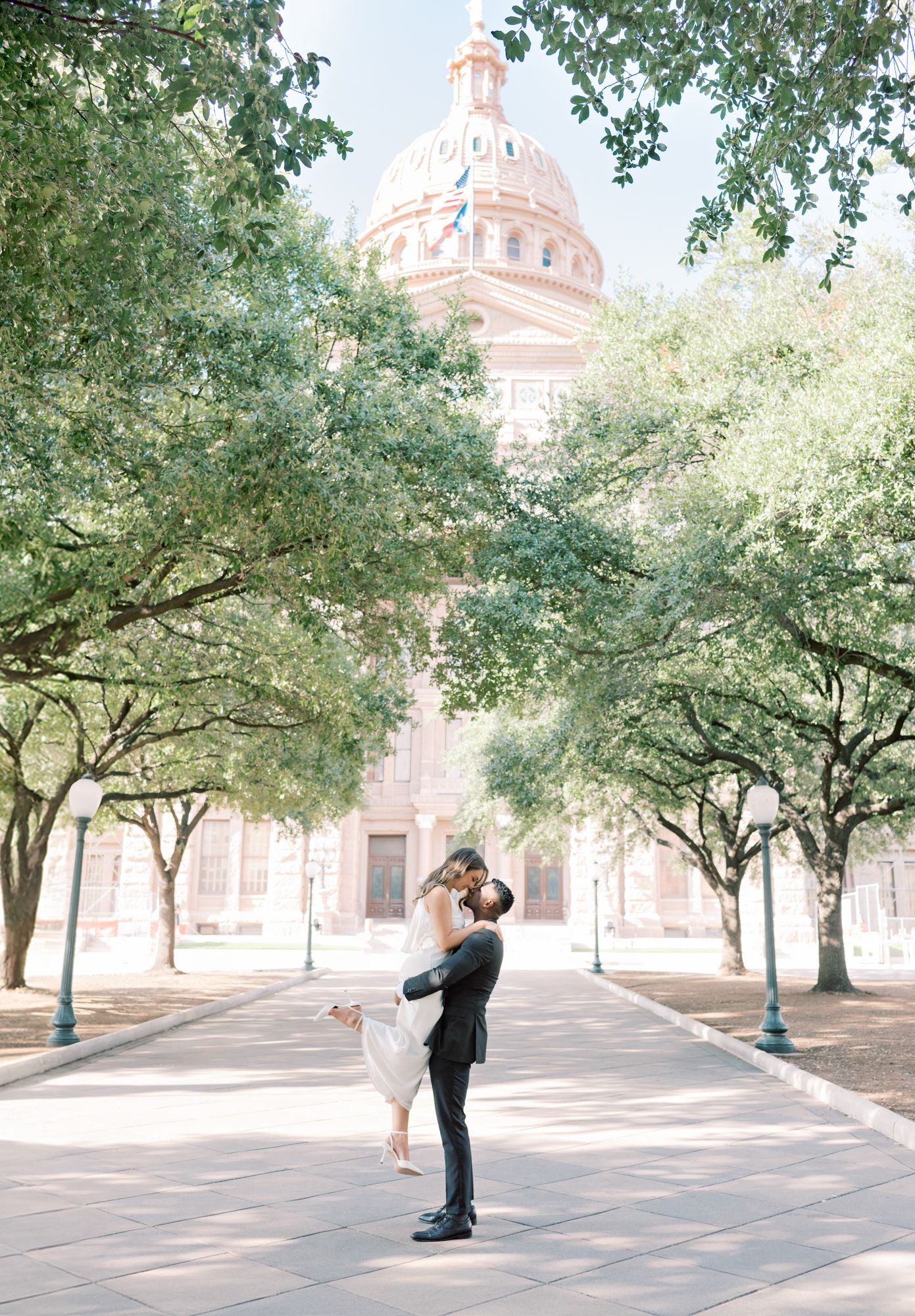 A couple embracing in front of a domed government building with tree-lined path.