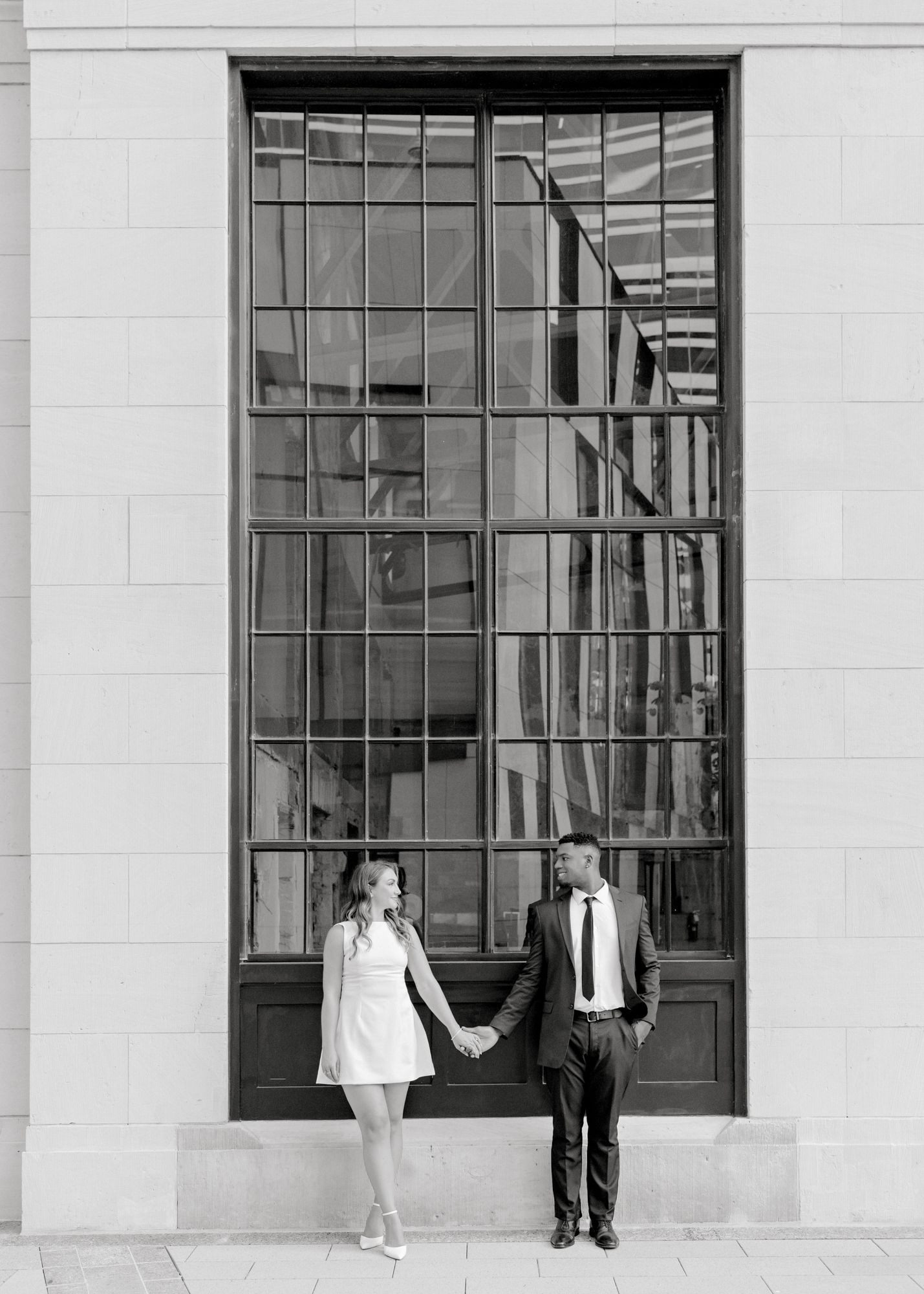 A couple standing in front of a tall window in black and white formal attire holding hands.