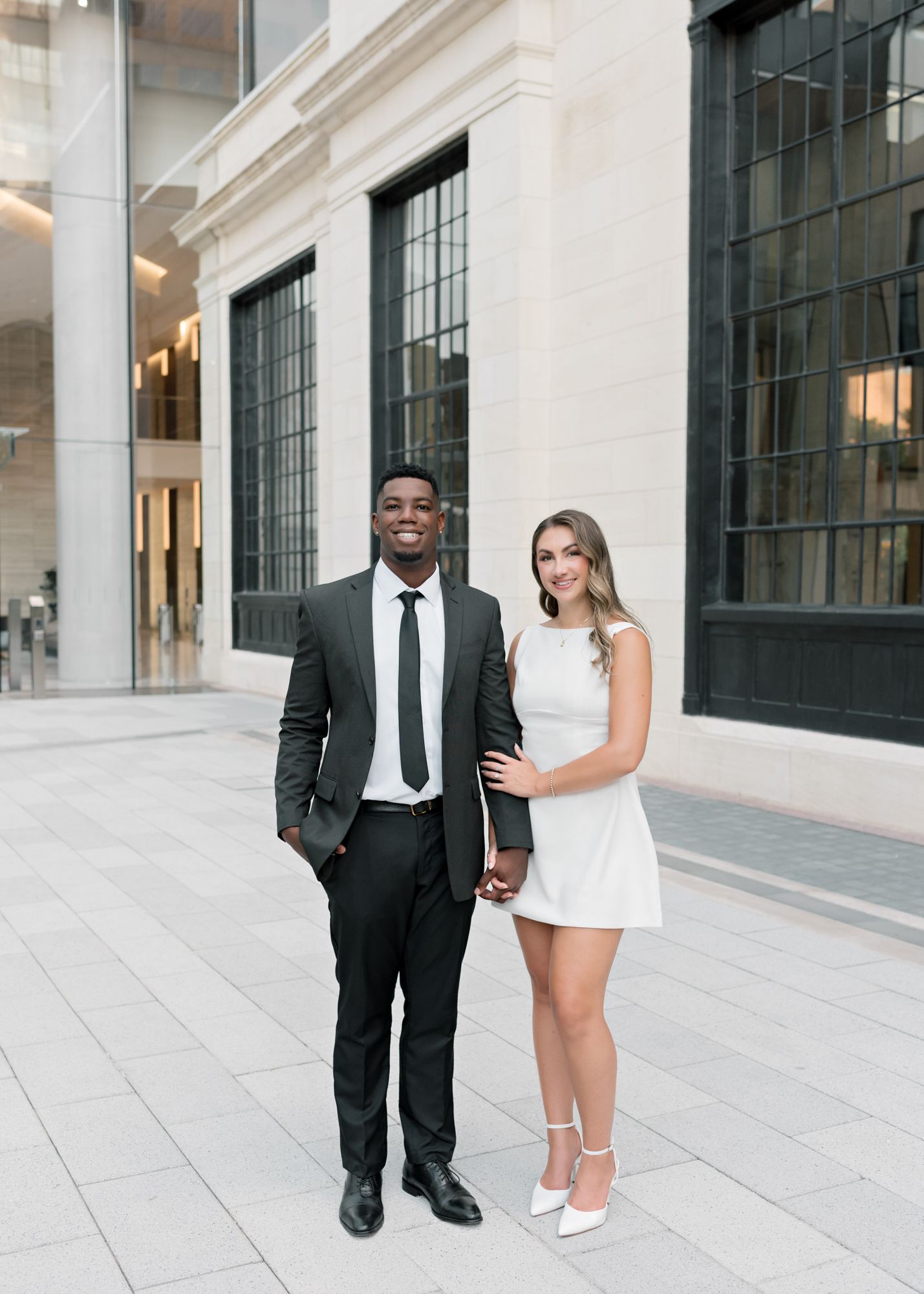 A couple poses together in formal attire against a modern building facade with large windows.