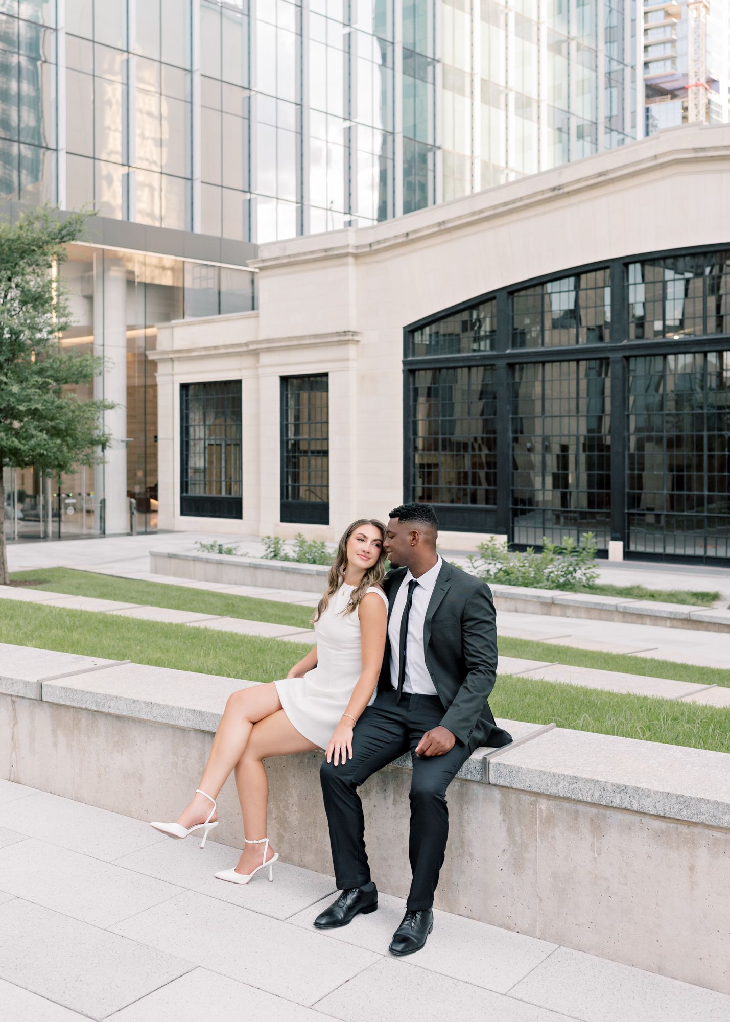A couple in formal attire sitting together on a concrete ledge outside a modern building.