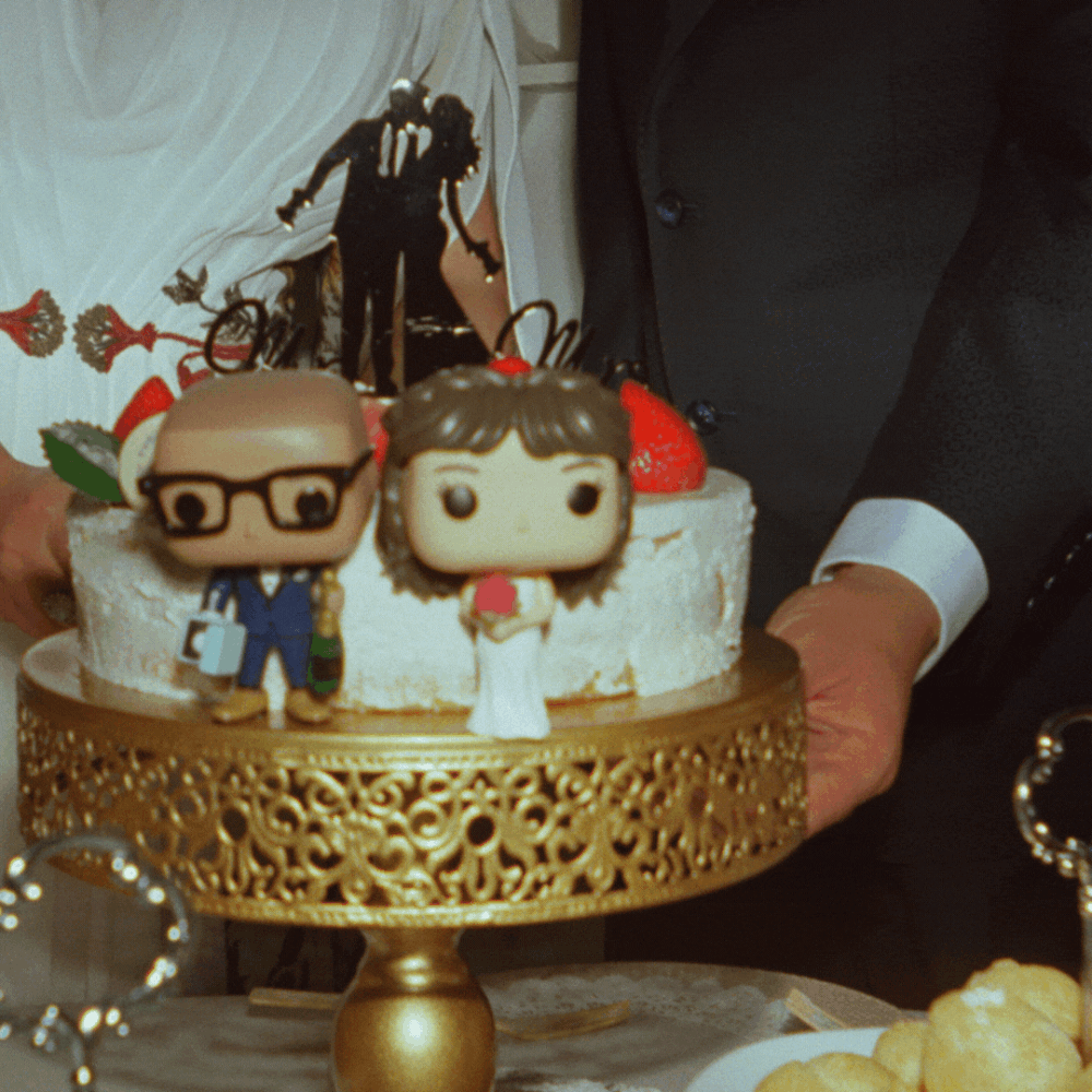 Wedding cake decorated with gold accents and Funko Pop style cake toppers displayed on an elegant gold stand.