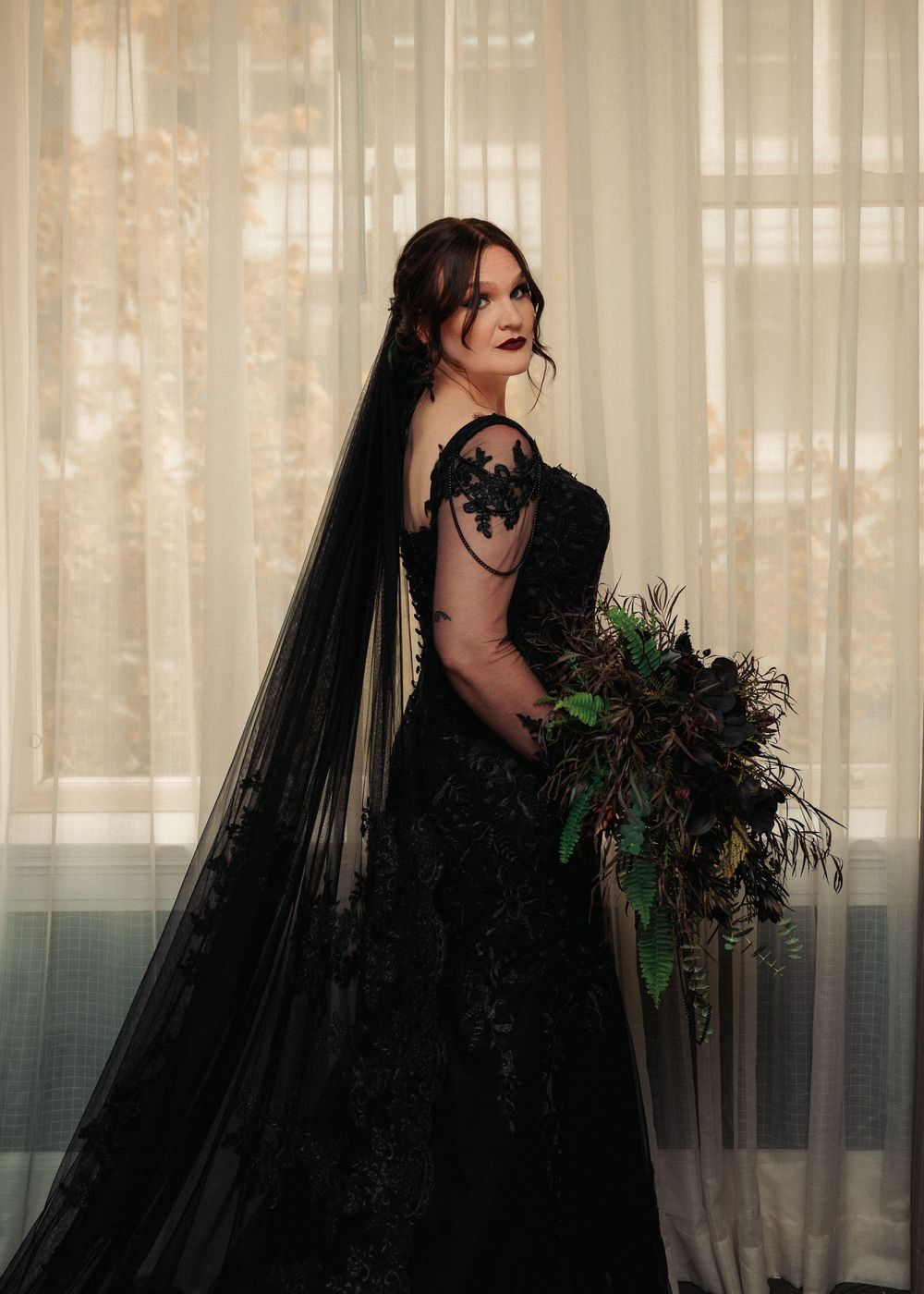 A dark-haired figure in black attire poses dramatically with a bouquet against a curtained window background.