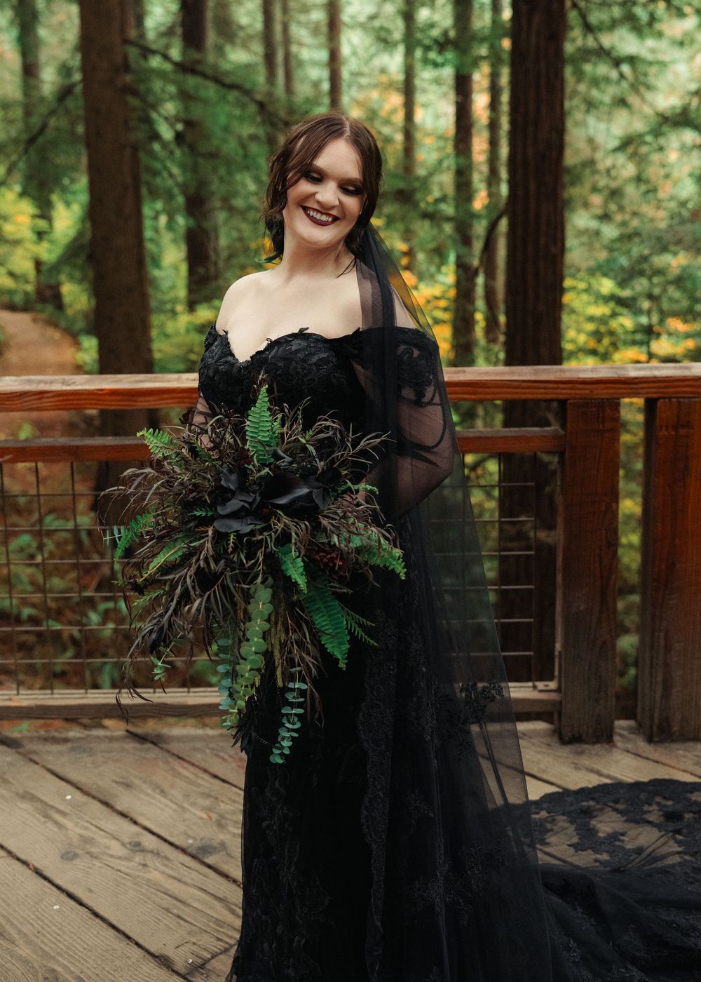 A black wedding dress with floral details photographed on a wooden deck in a forest setting.