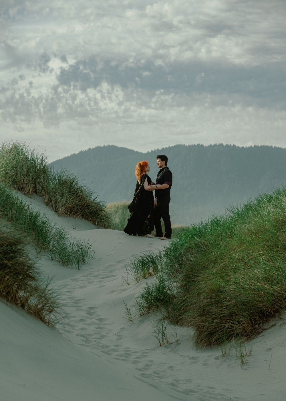 Couple in black attire embracing on sandy dunes with green grass and misty mountains under cloudy skies.