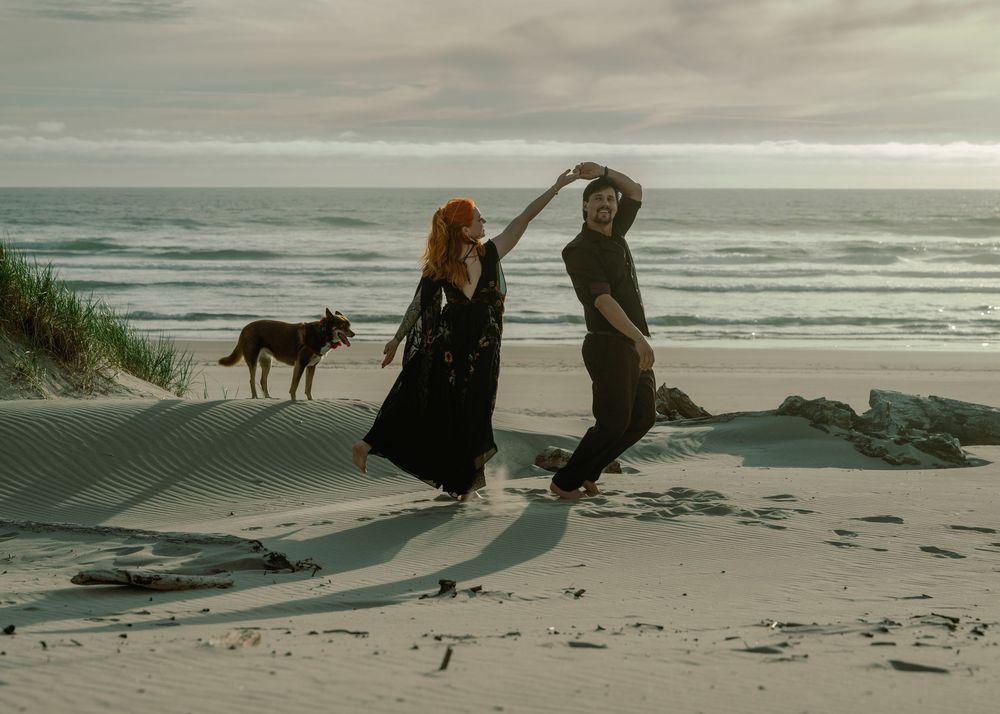 Couple dancing on a sandy beach with a dog and ocean waves in the background on a cloudy day.