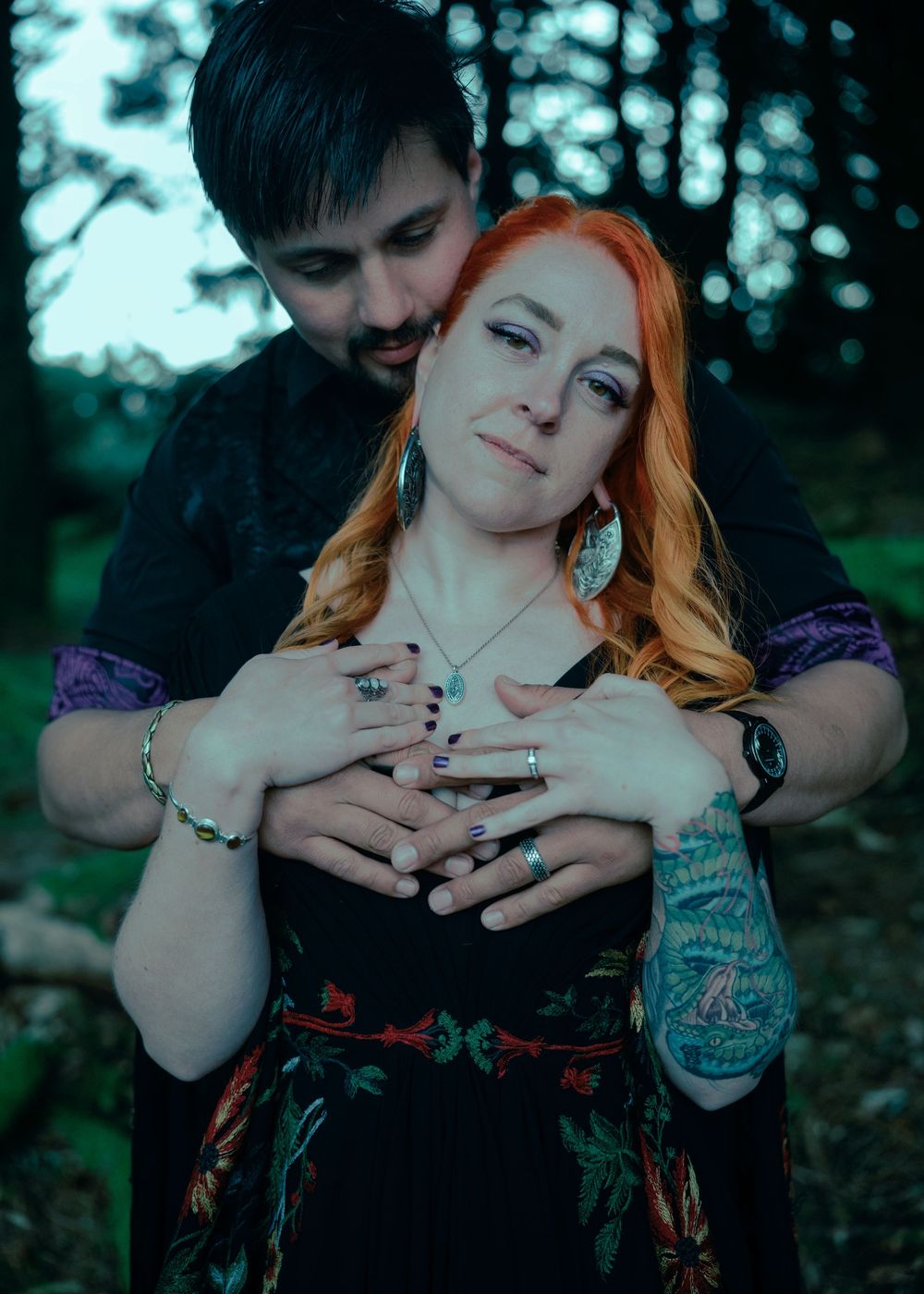 A couple embraces in a forest, woman with red hair wearing jewelry and a floral dress, man with tattoos behind her.