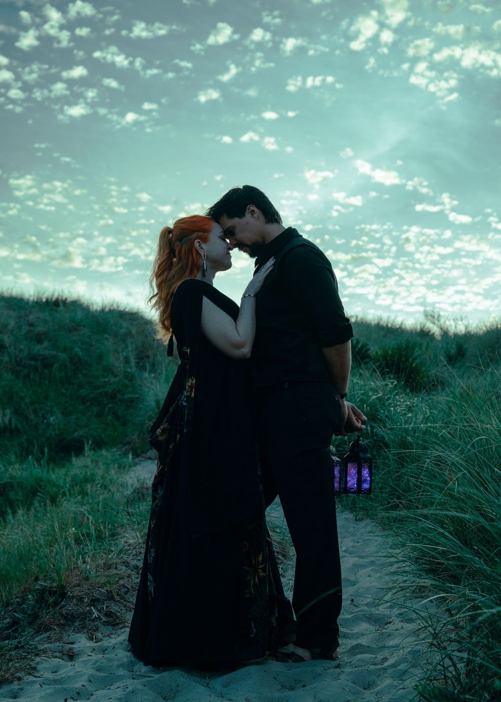 Romantic couple in black attire sharing an intimate moment on a sandy path surrounded by green dunes under a dramatic teal sky.
