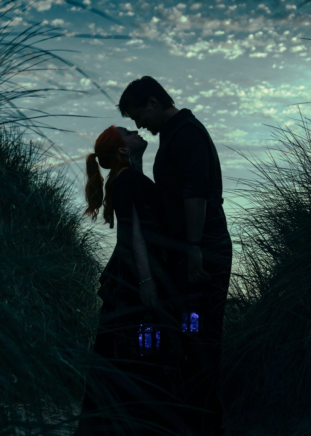 Romantic couple silhouetted against a dramatic twilight sky, sharing an intimate moment among coastal dunes at dusk.