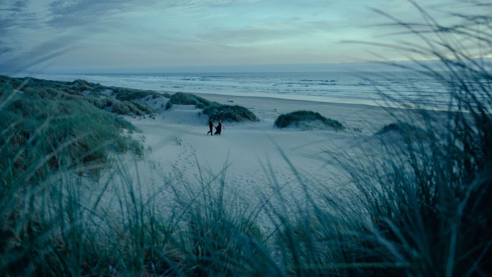 Two people stand on a serene sandy beach at dusk, surrounded by dune grass with ocean waves in the background.