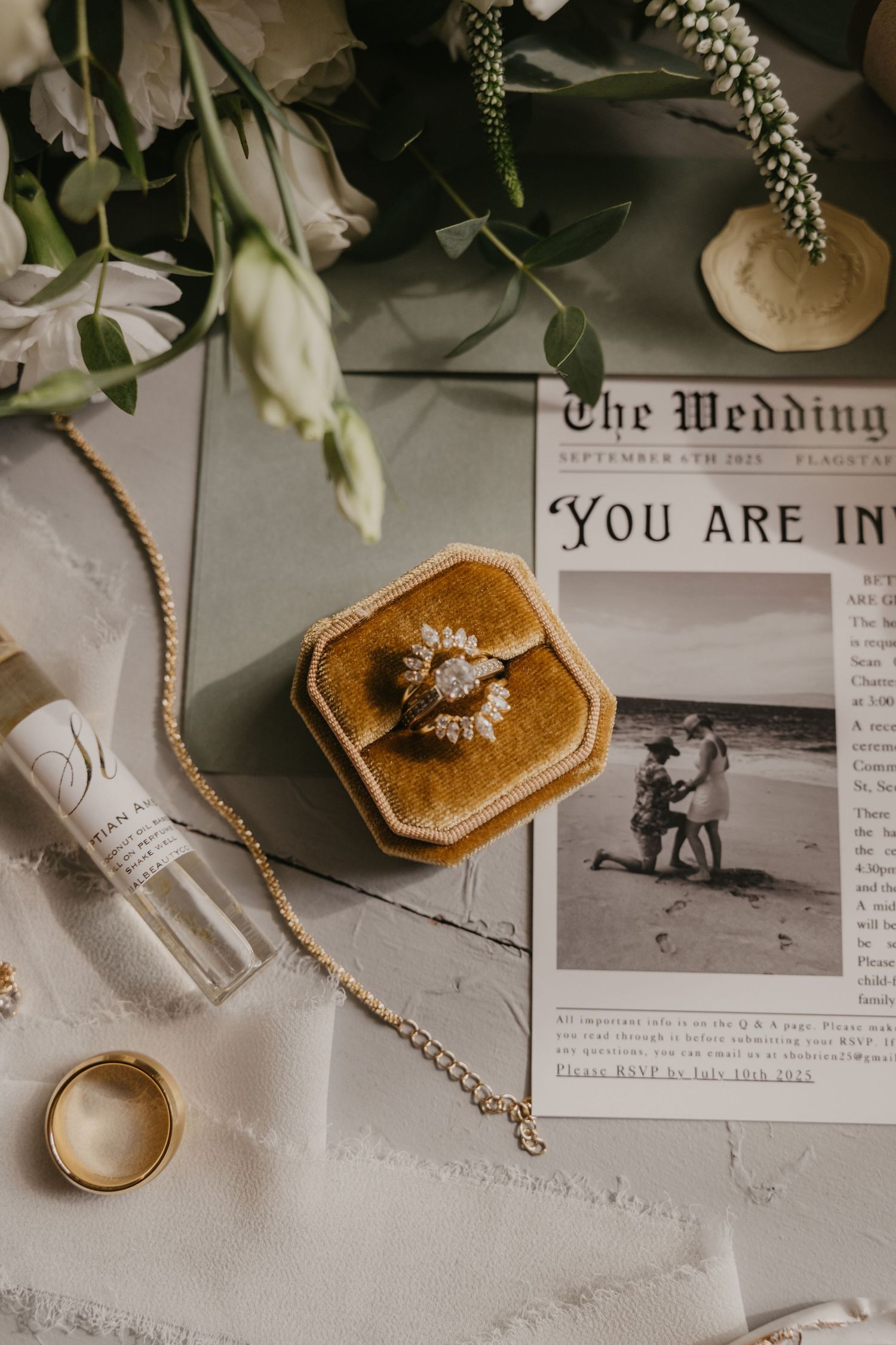 Wedding detail flatlay with rings, dried flowers, and vintage newspaper clippings on white textured surface.