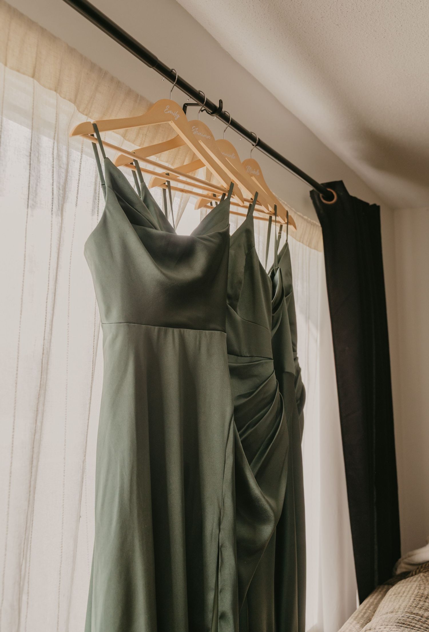 Sage green bridesmaid dresses hanging on wooden hangers against white curtains.