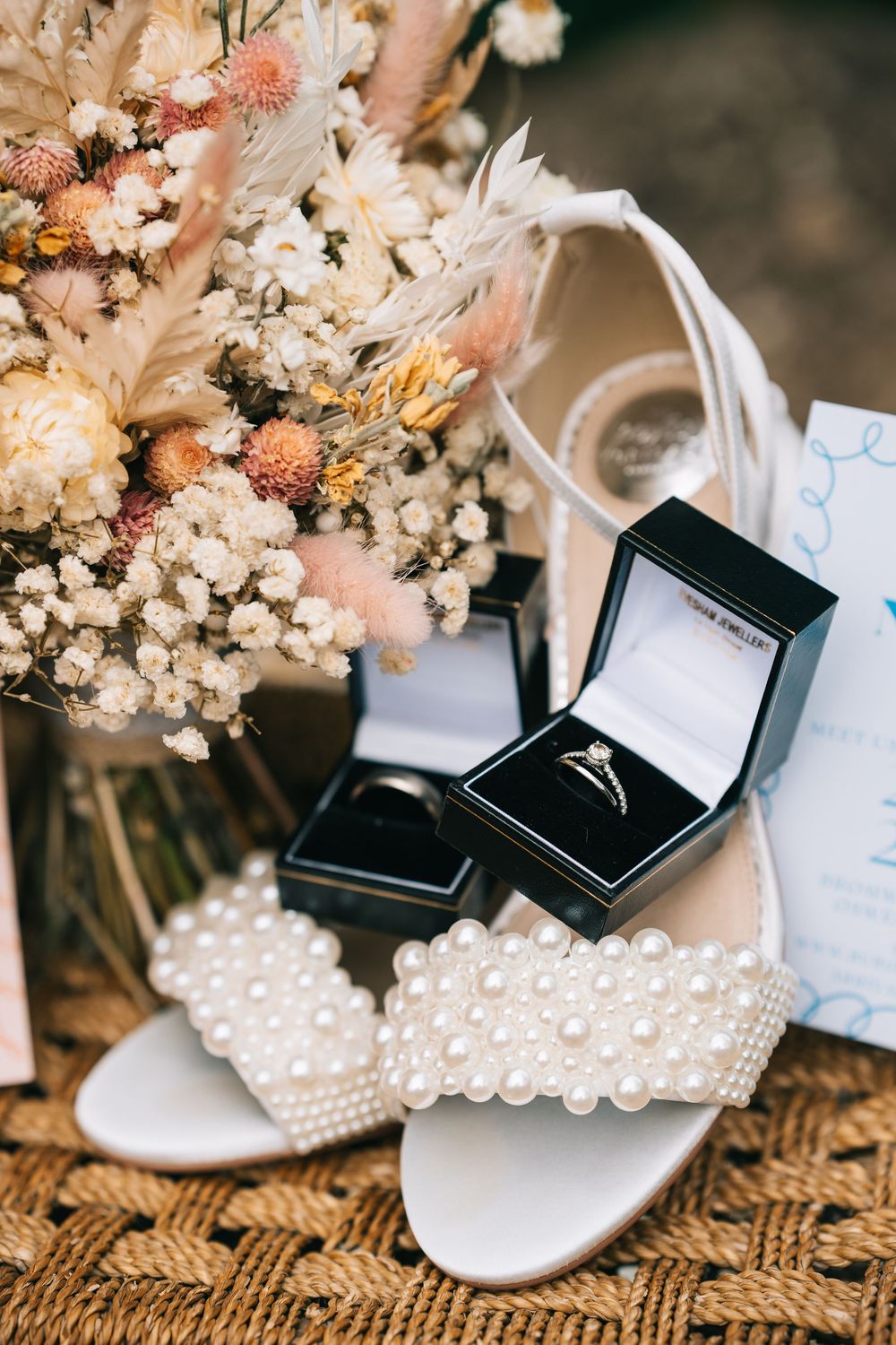Wedding accessories including pearl-adorned bridal shoes and black ring boxes arranged with dried flowers on textured fabric.