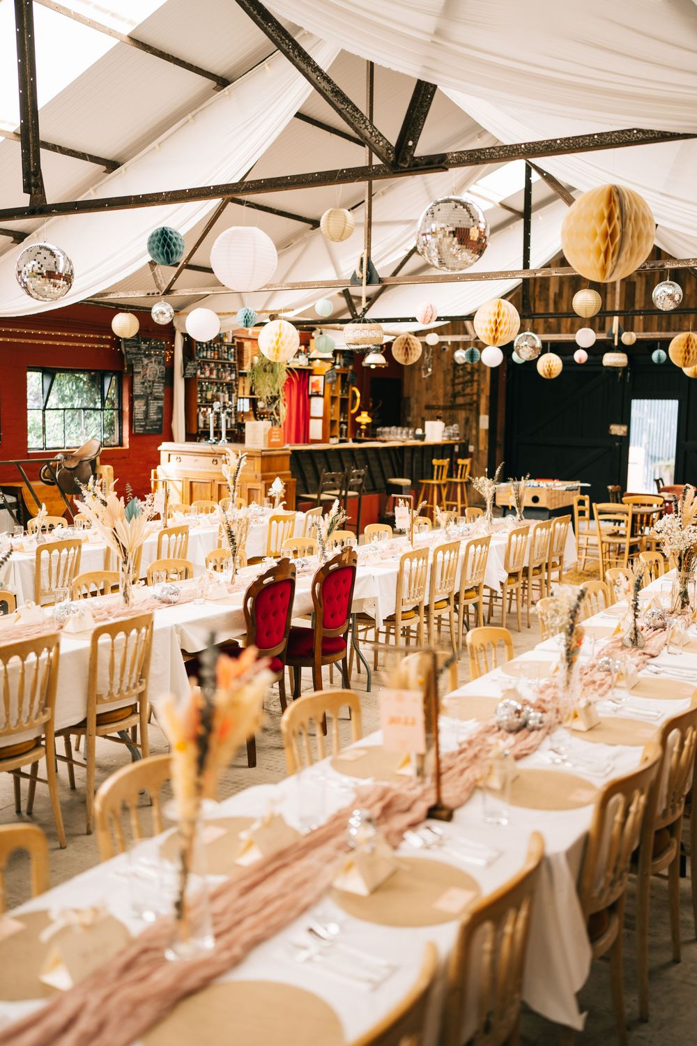 Long banquet tables set with white linens and chairs under a tent with hanging string lights and disco balls.