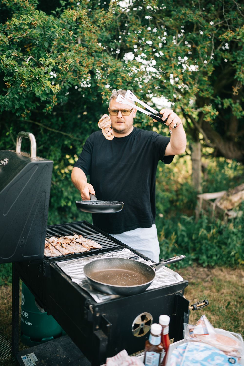 Someone grilling food outdoors on a barbecue grill with trees in the background during a sunny day.