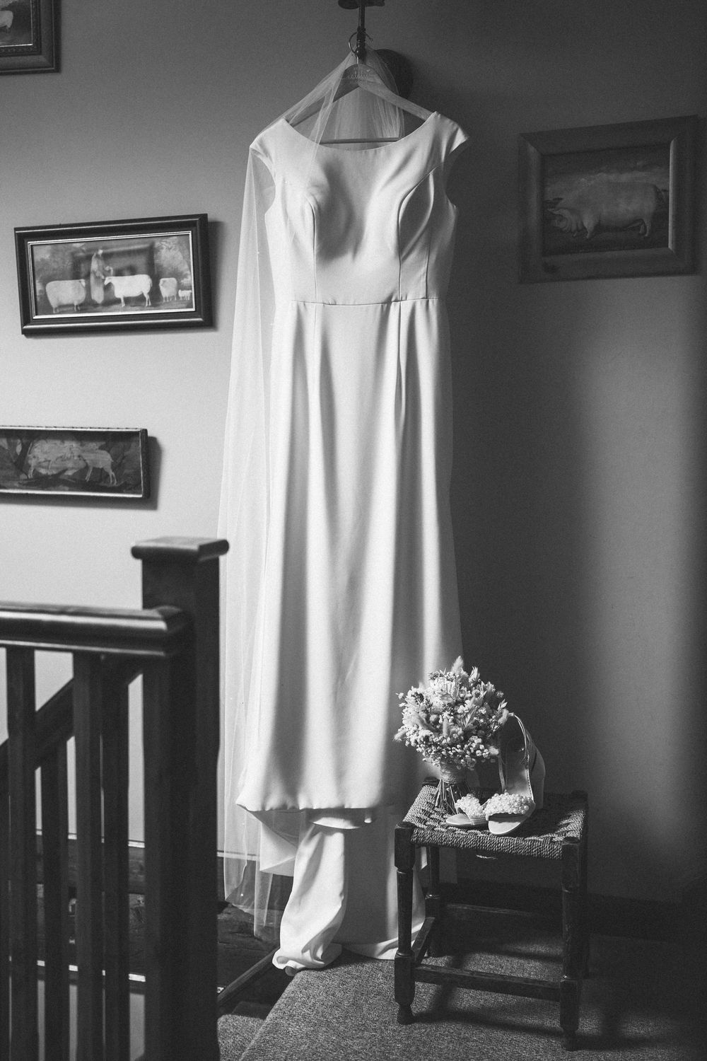 A white wedding dress hanging on display in a dark room with wooden furniture and framed artwork on the walls.