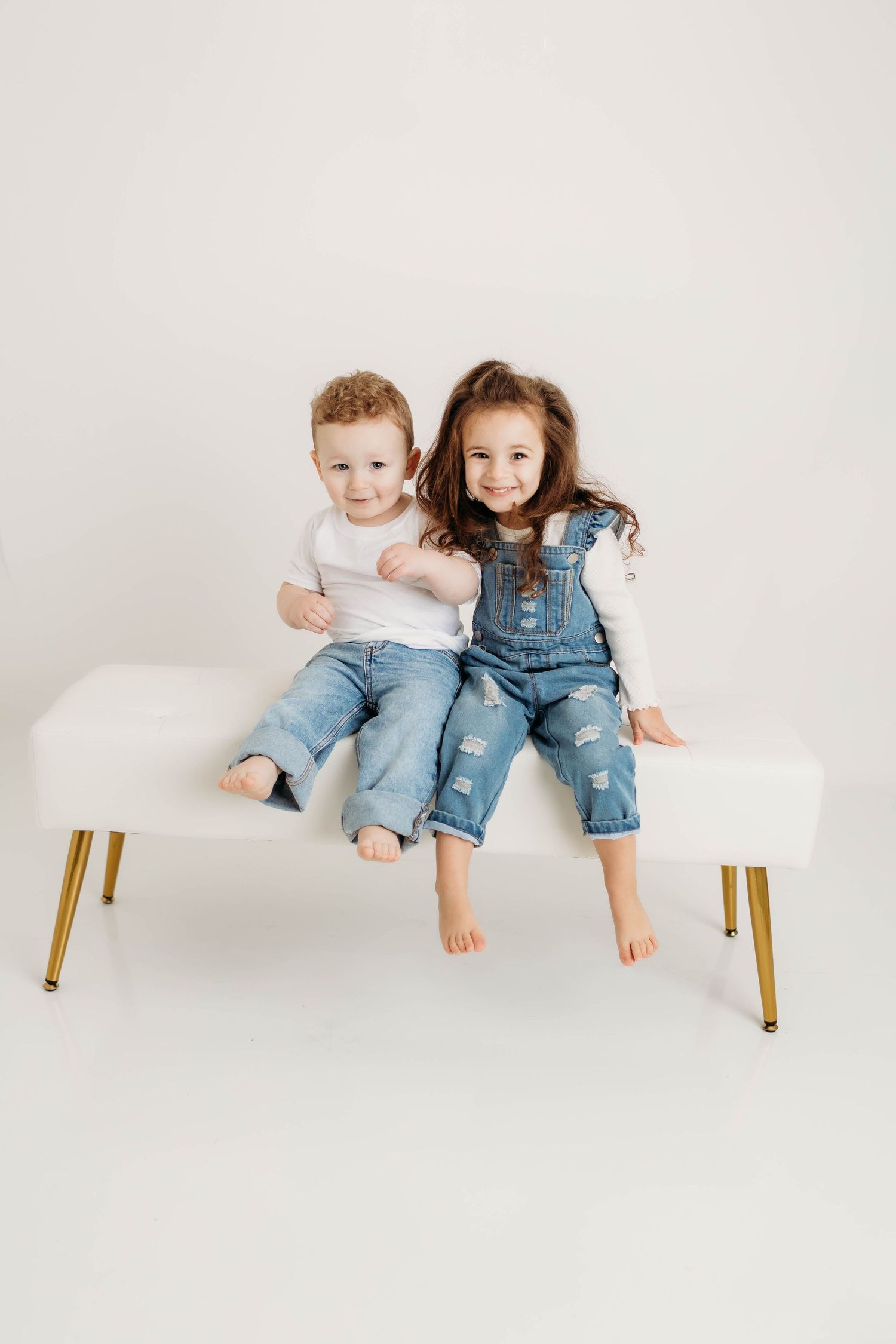 Two young children in denim sit barefoot on a white bench with gold legs against a light background.