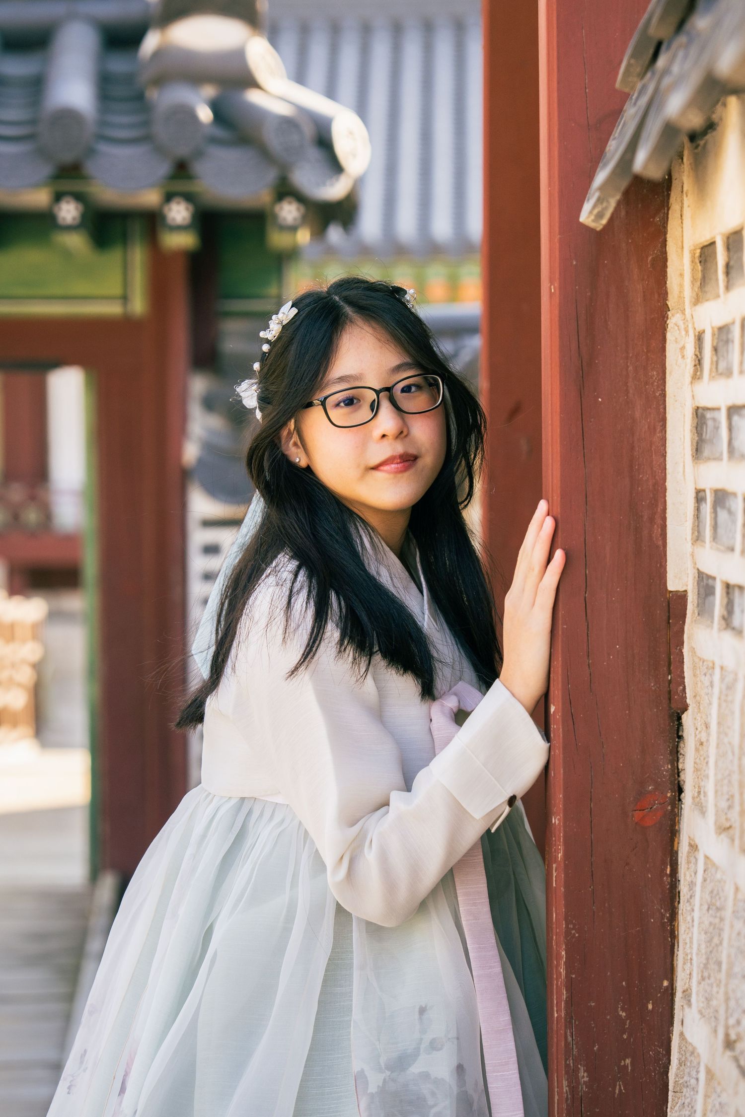 A person wearing traditional Korean hanbok dress and glasses stands by a temple wall in soft natural lighting.