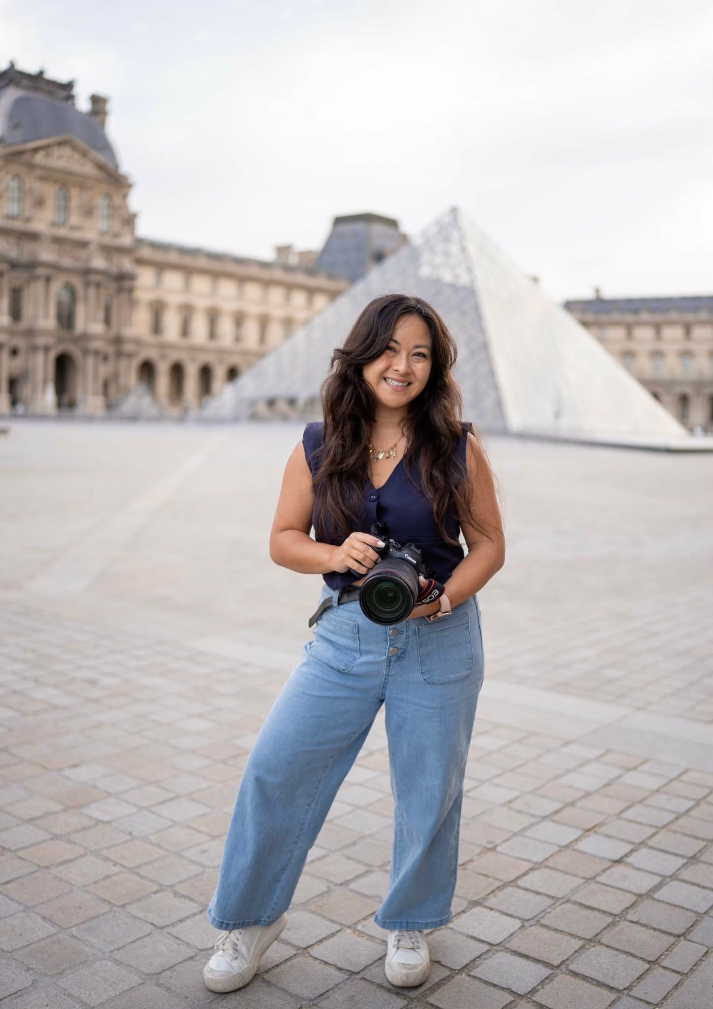 Tourist with camera poses in front of the iconic glass pyramid at the Louvre Museum in Paris wearing casual denim.