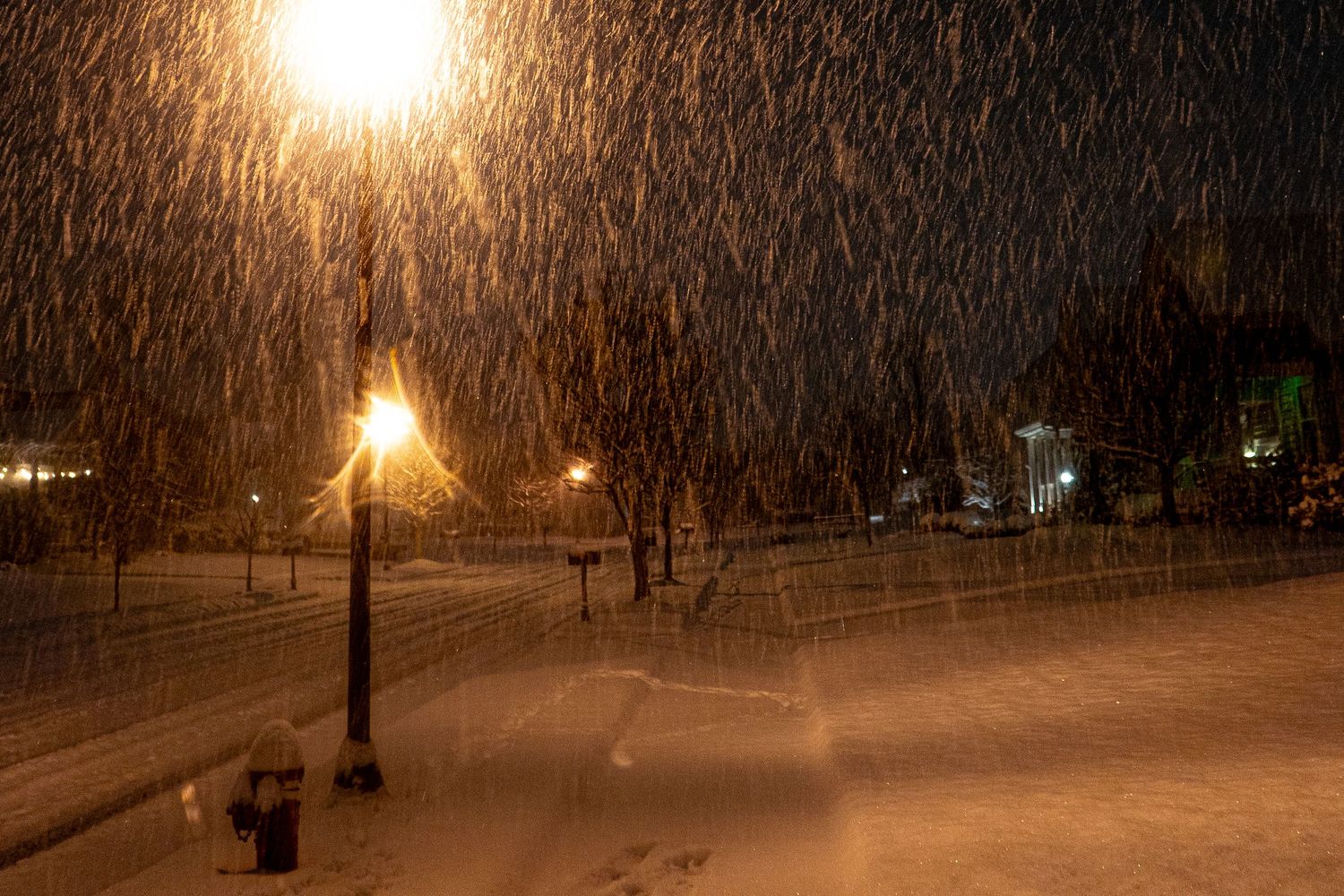 Snow falls heavily at night illuminated by warm street lights casting an ethereal glow over the winter scene.