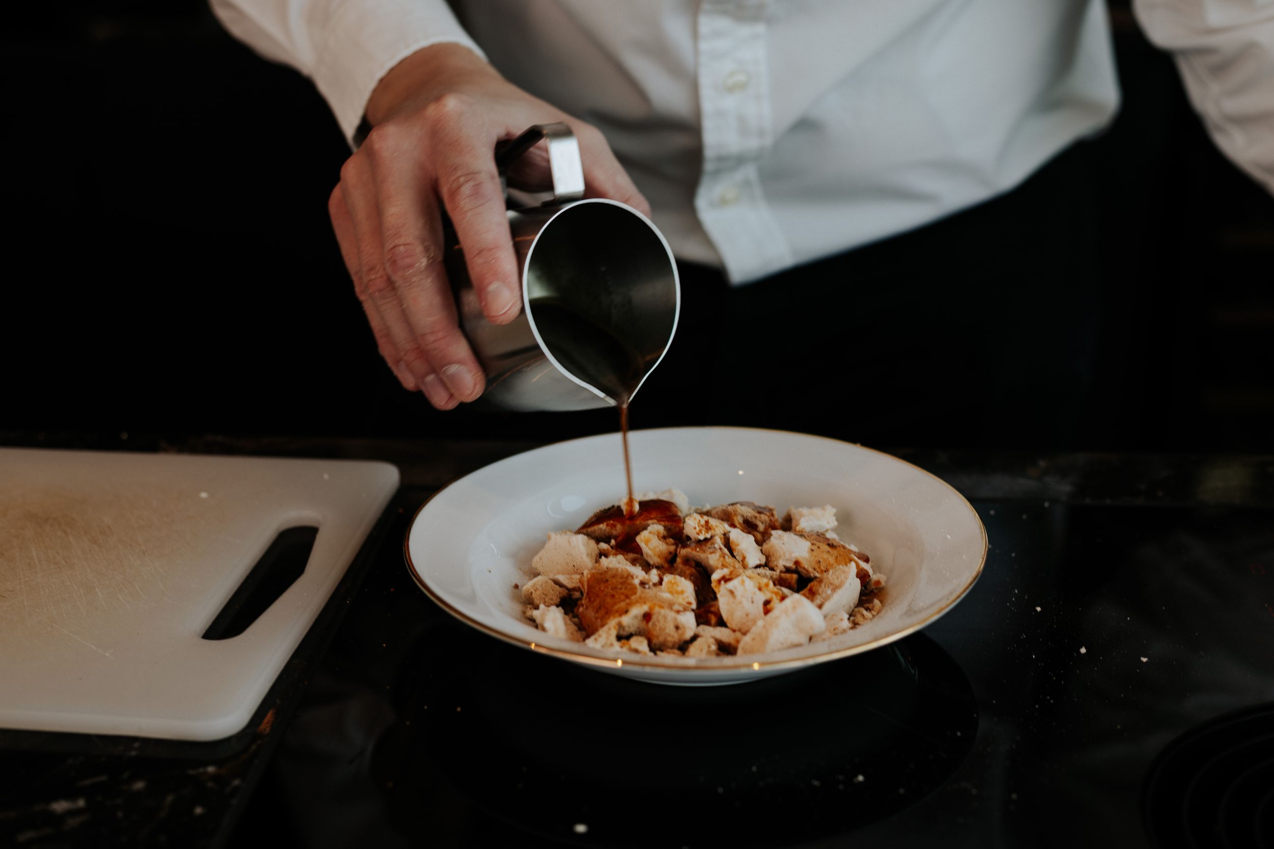 Hands sprinkling toppings onto a white plate of food in a dimly lit setting.