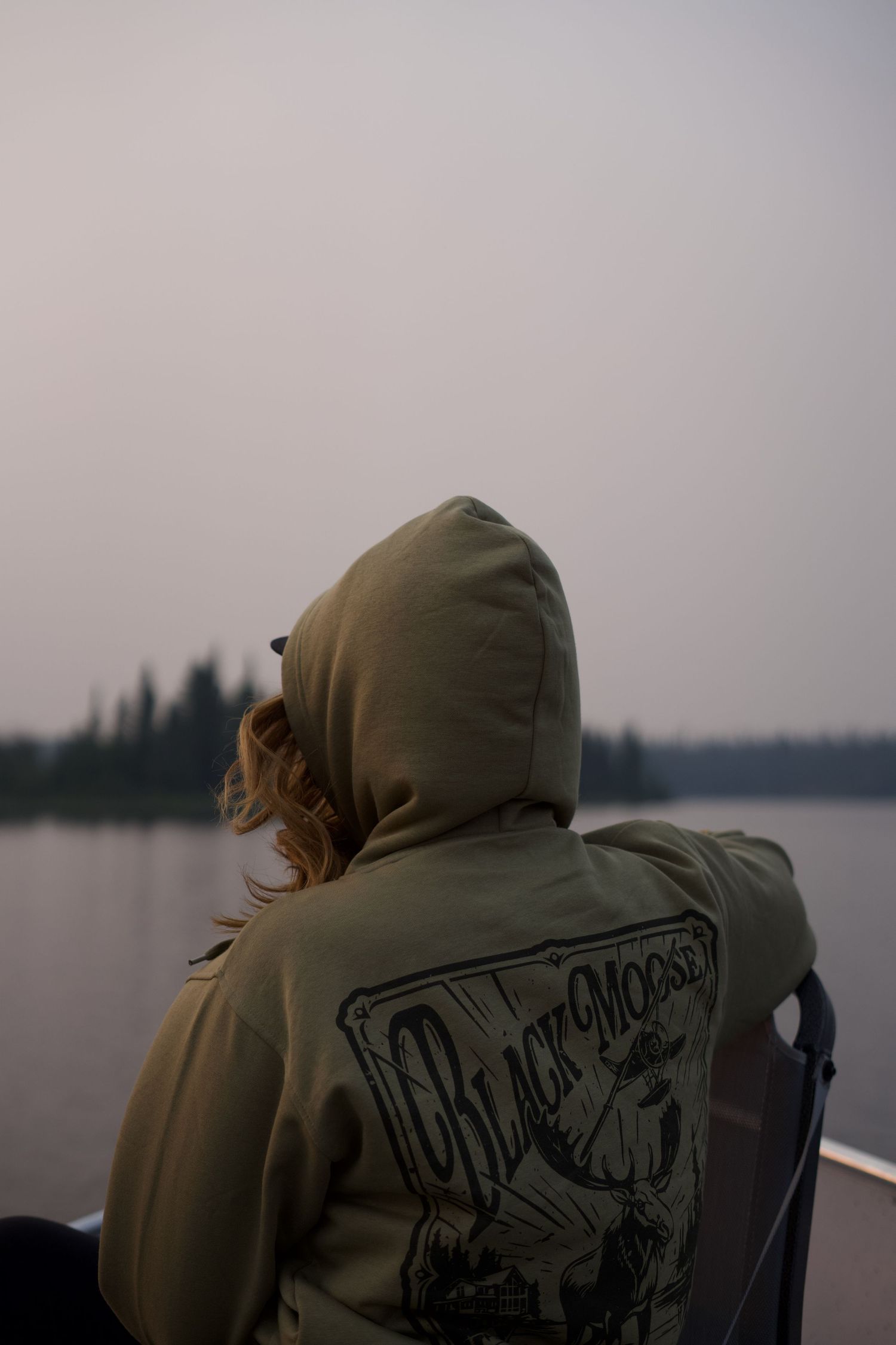 A person in a hooded jacket sits looking out over a foggy lake at dusk.