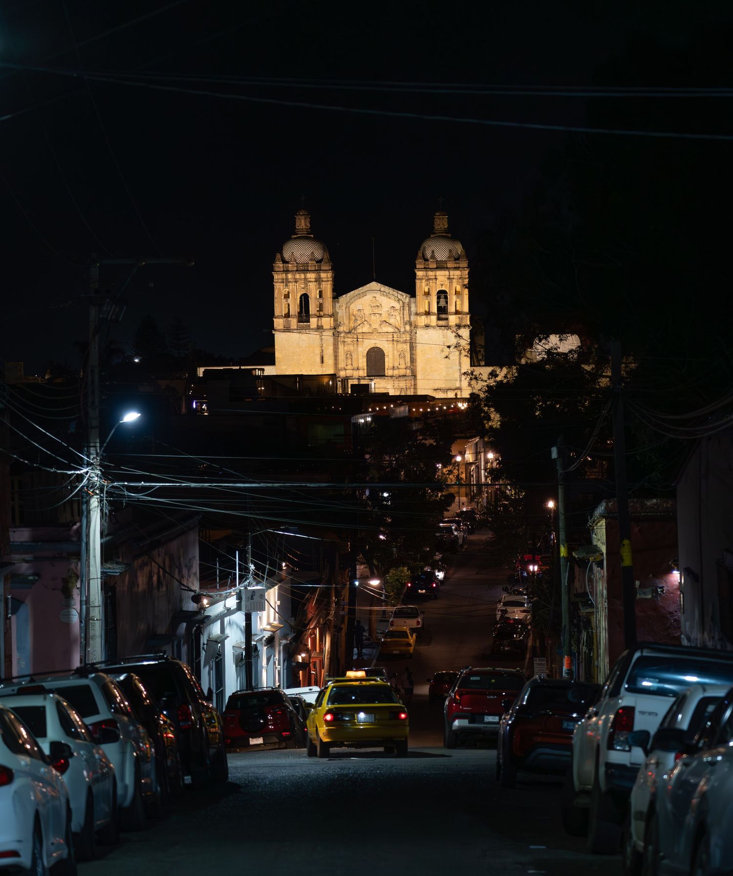 Templo de Santo Domingo church illuminated at night with parked cars along a dark street in Oaxaca, Mexico.