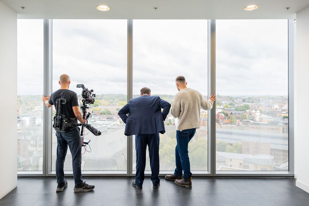 Film crew working near large windows overlooking cityscape in modern office building.