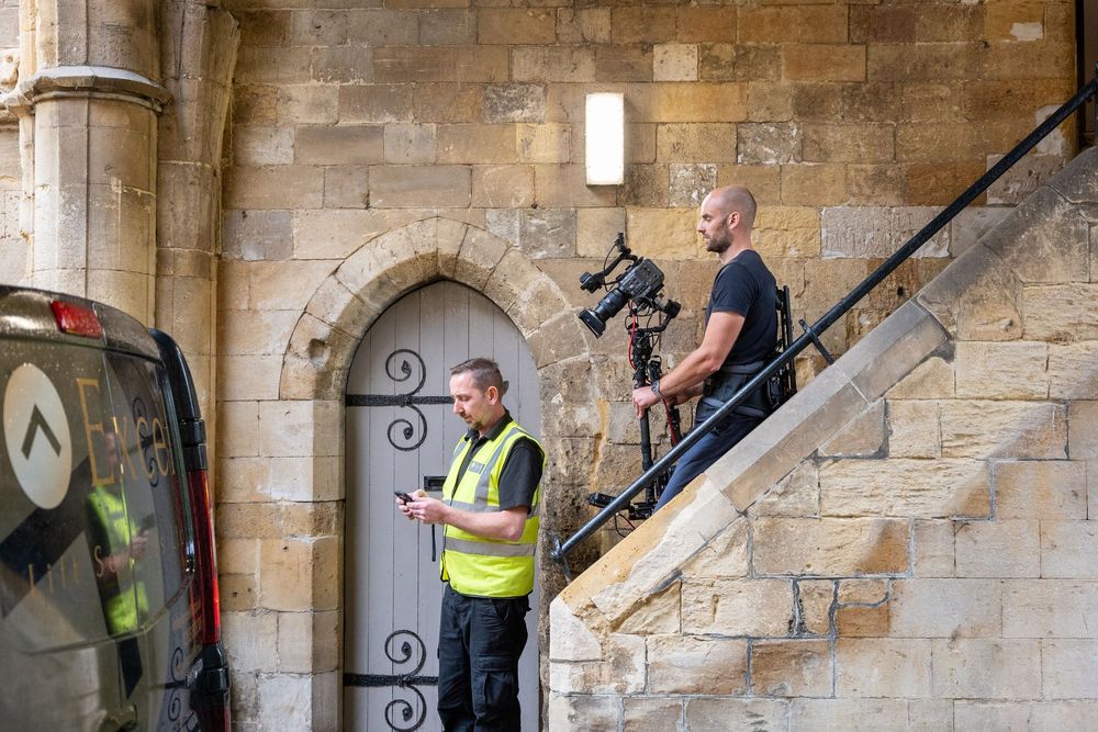 Workers carry a large piece of furniture up a narrow stone staircase in a historic building.