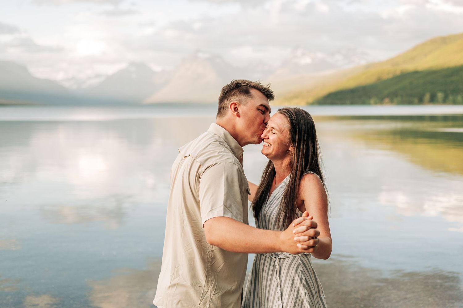 Romantic couple embraces at sunset by a serene mountain lake with golden light reflecting on the water.