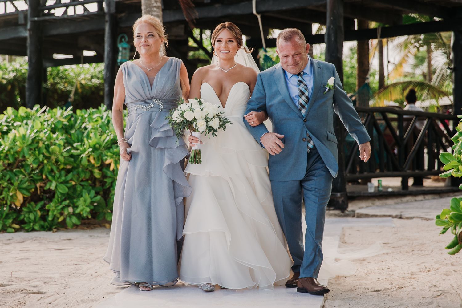 Three people walk arm in arm on a garden path, with the person in the middle wearing a white wedding gown. El Dorado Royale Wedding Photo