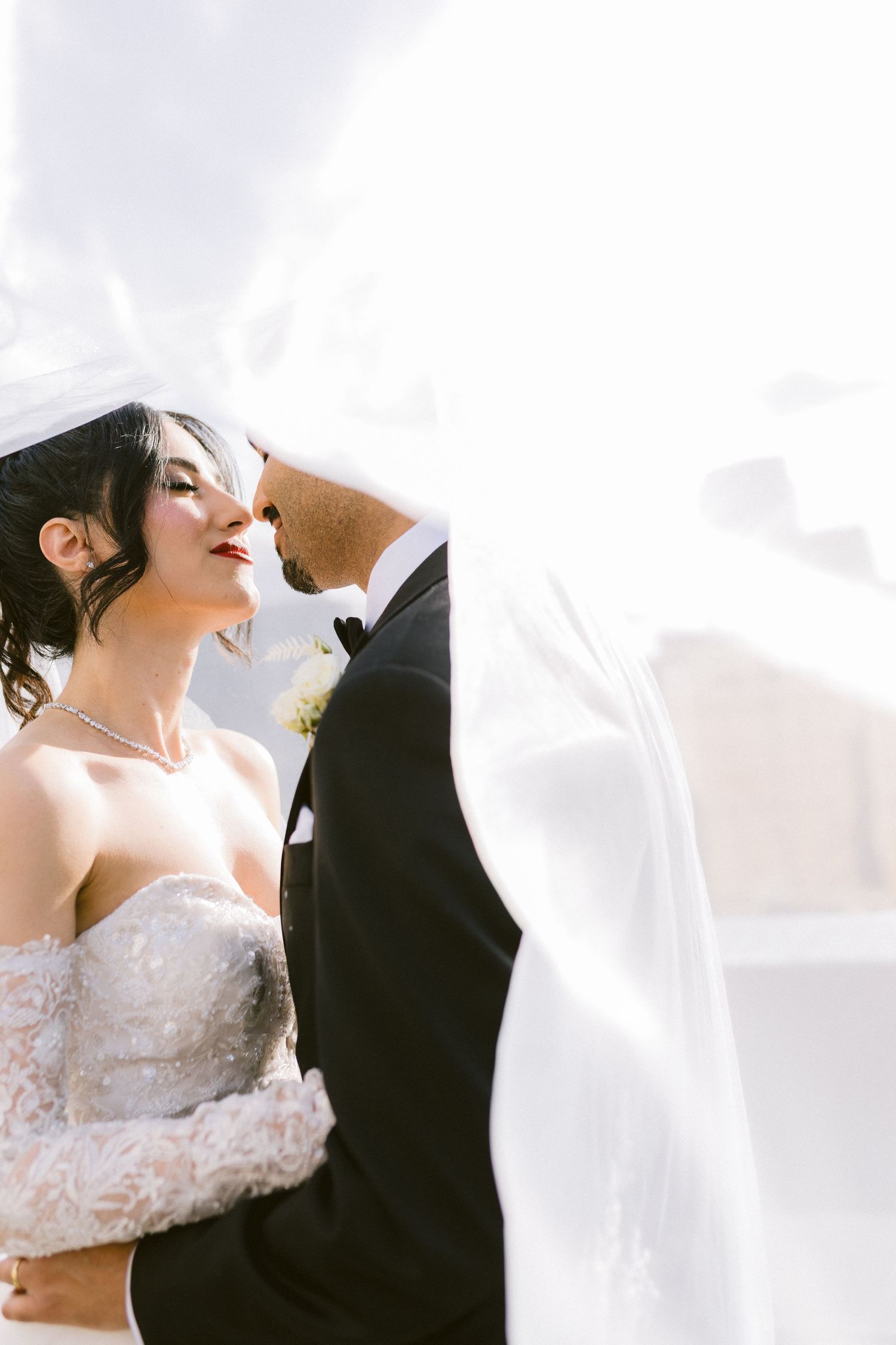 Romantic wedding couple embraces under flowing white veil against bright sky in artistic composition.