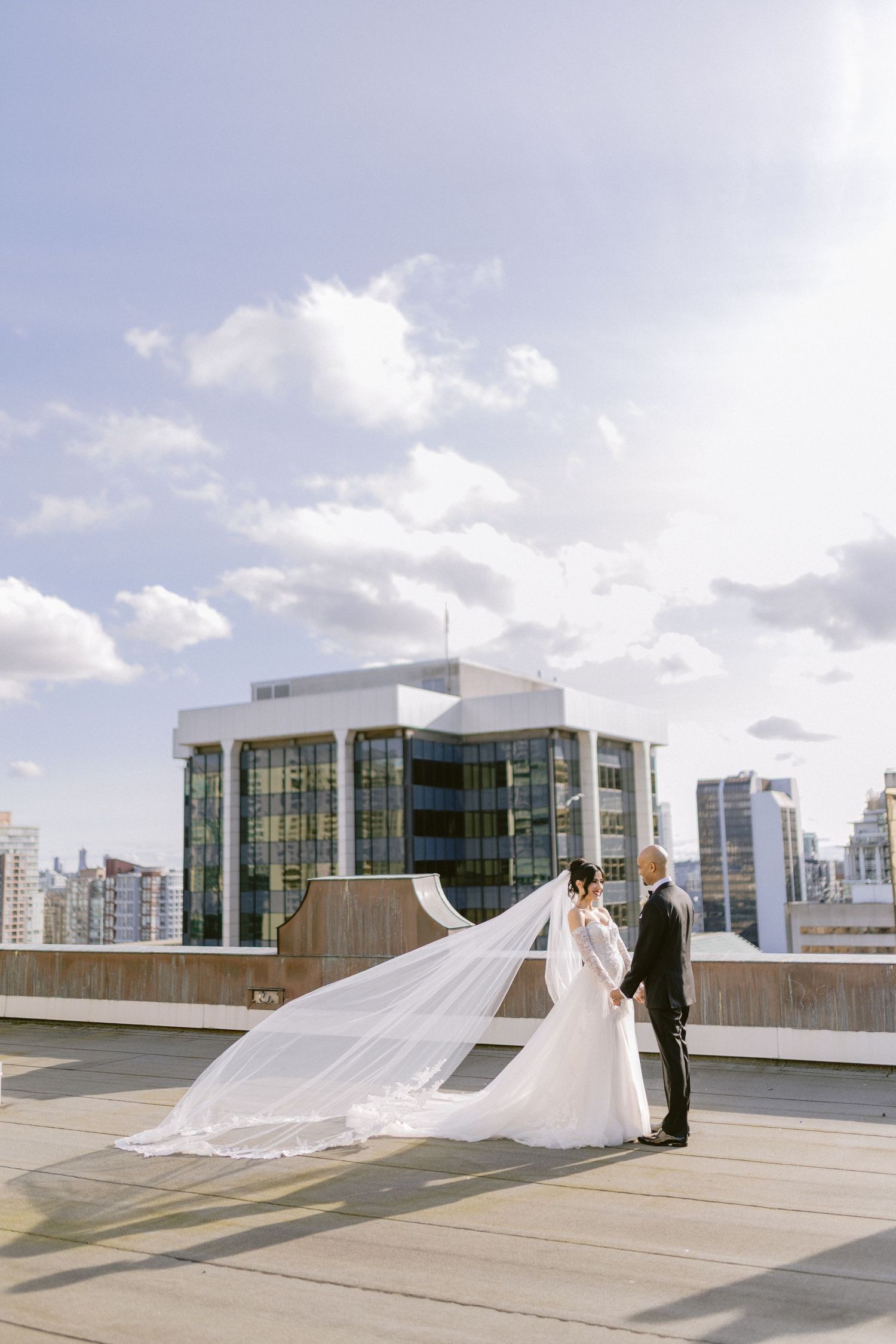 A wedding couple poses on a modern rooftop terrace with a dramatic flowing veil and city skyline in the background.