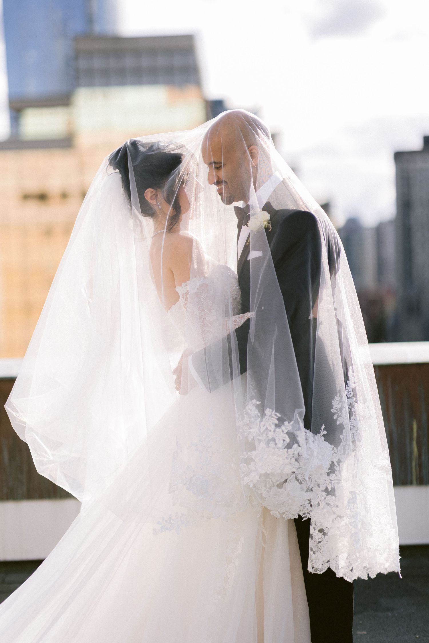 Romantic wedding couple embraces under flowing white lace veil against city skyline backdrop.