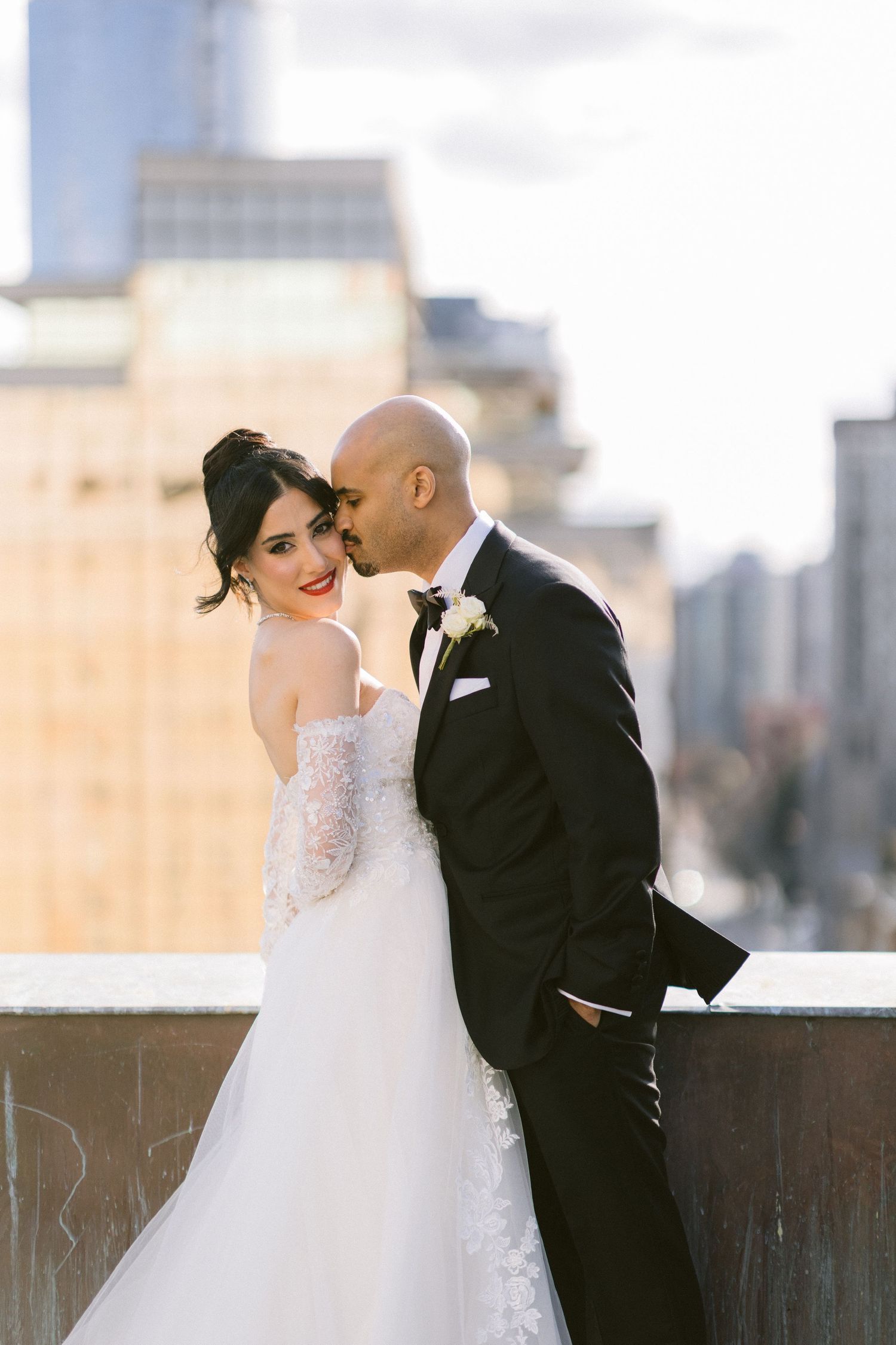A romantic wedding couple embraces on a rooftop with a cityscape backdrop at sunset.