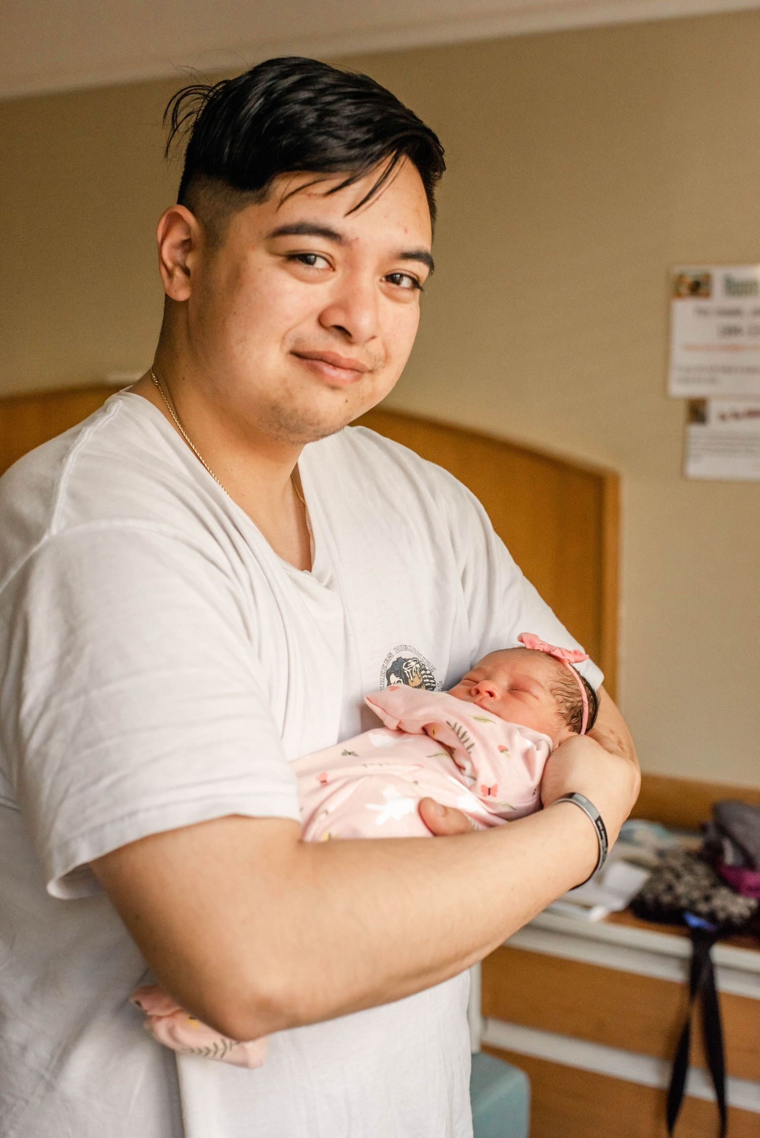 Parent in white t-shirt holding and gazing lovingly at newborn baby wrapped in pink blanket in hospital room.