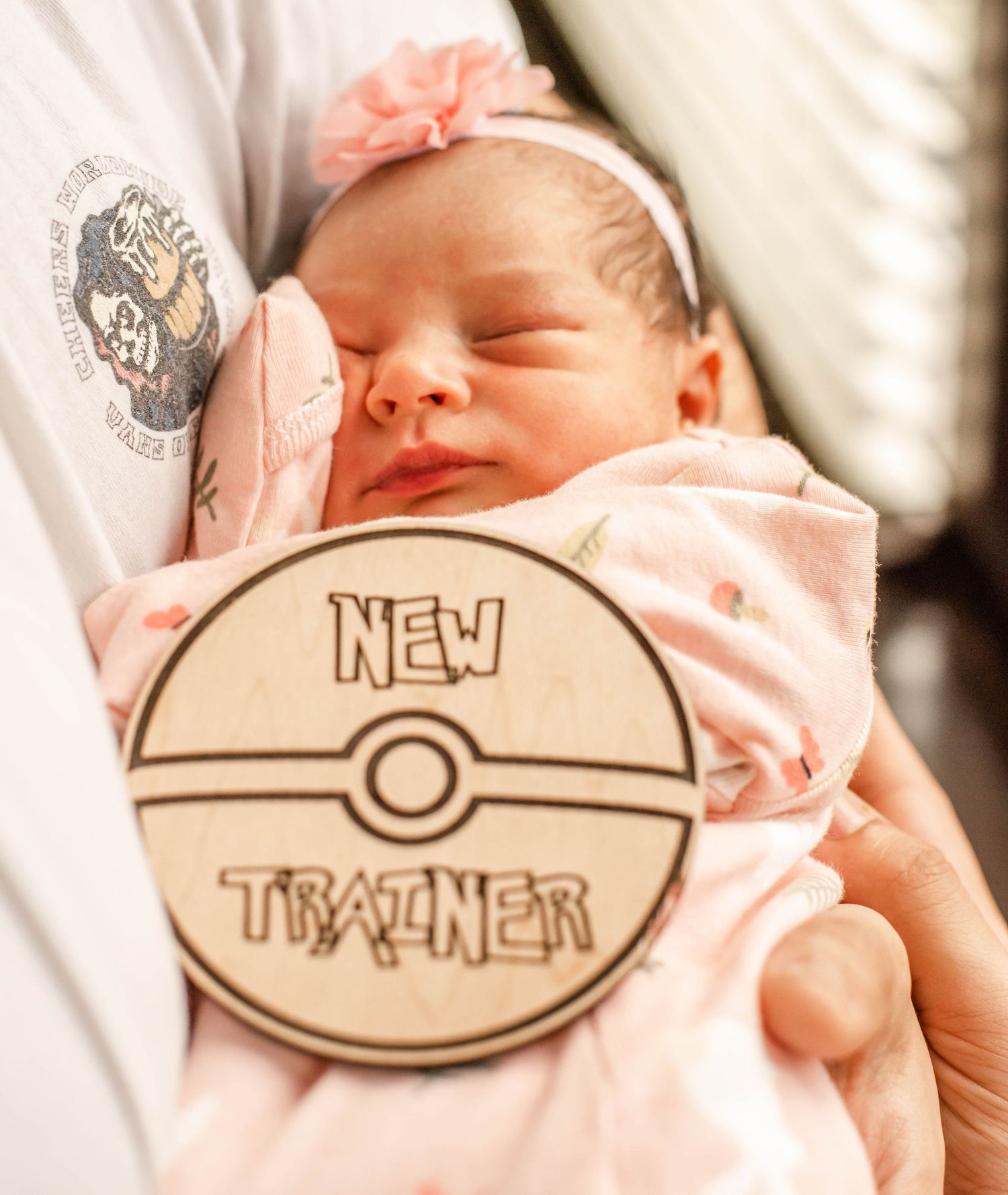 A newborn baby sleeps peacefully in white bedding next to a wooden Pokemon-themed 'New Trainer' sign.