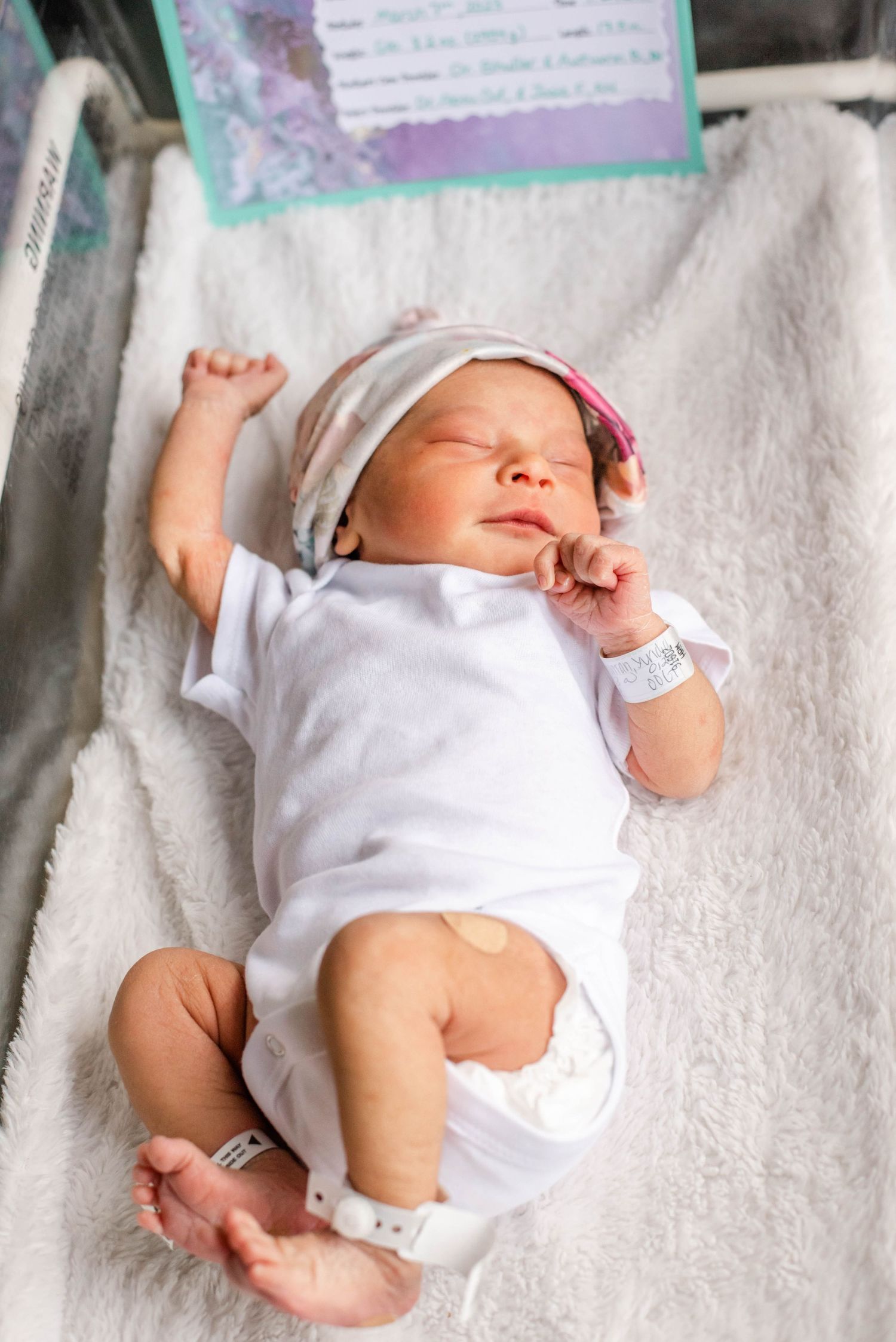A sleeping newborn baby wearing a white onesie lies peacefully on soft white bedding.
