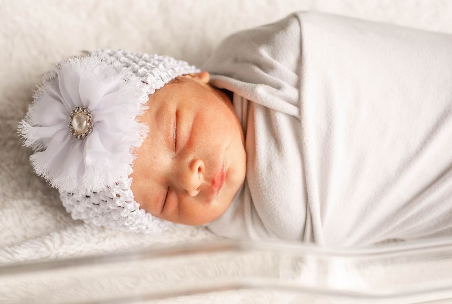 Sleeping baby wrapped in white blanket with delicate lace headband lying on white bedding.