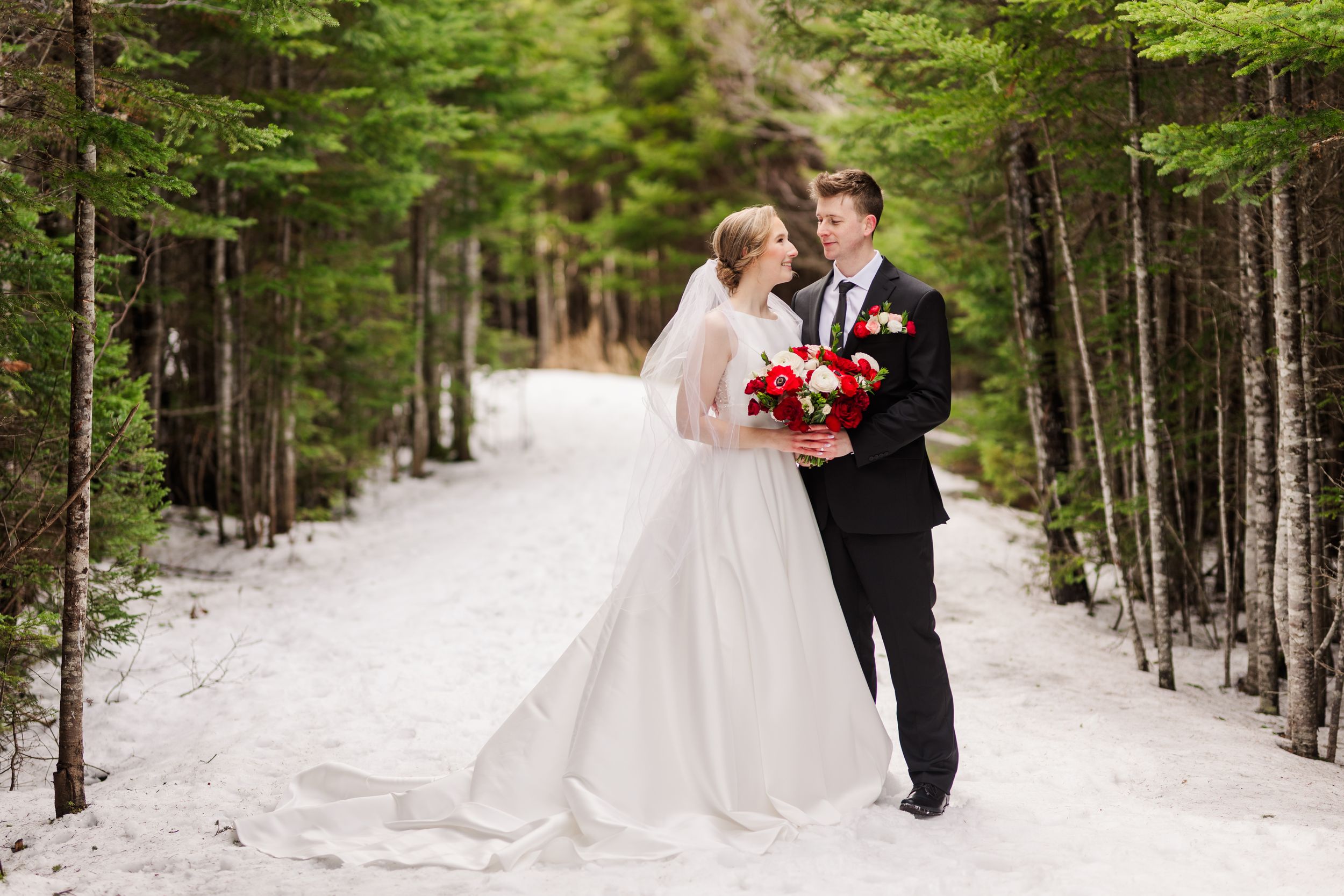 A romantic winter wedding couple walks through a snow-covered forest path lined with evergreen trees.