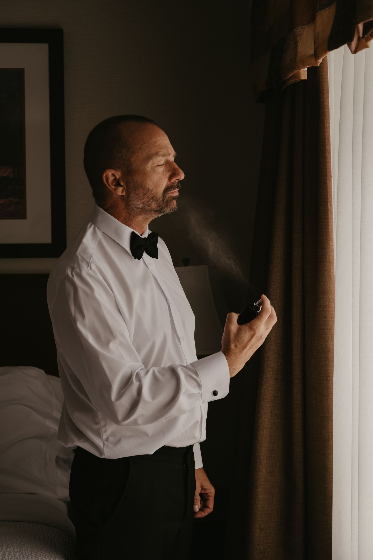 A person in a white dress shirt and black bow tie stands in a dimly lit room with curtains.