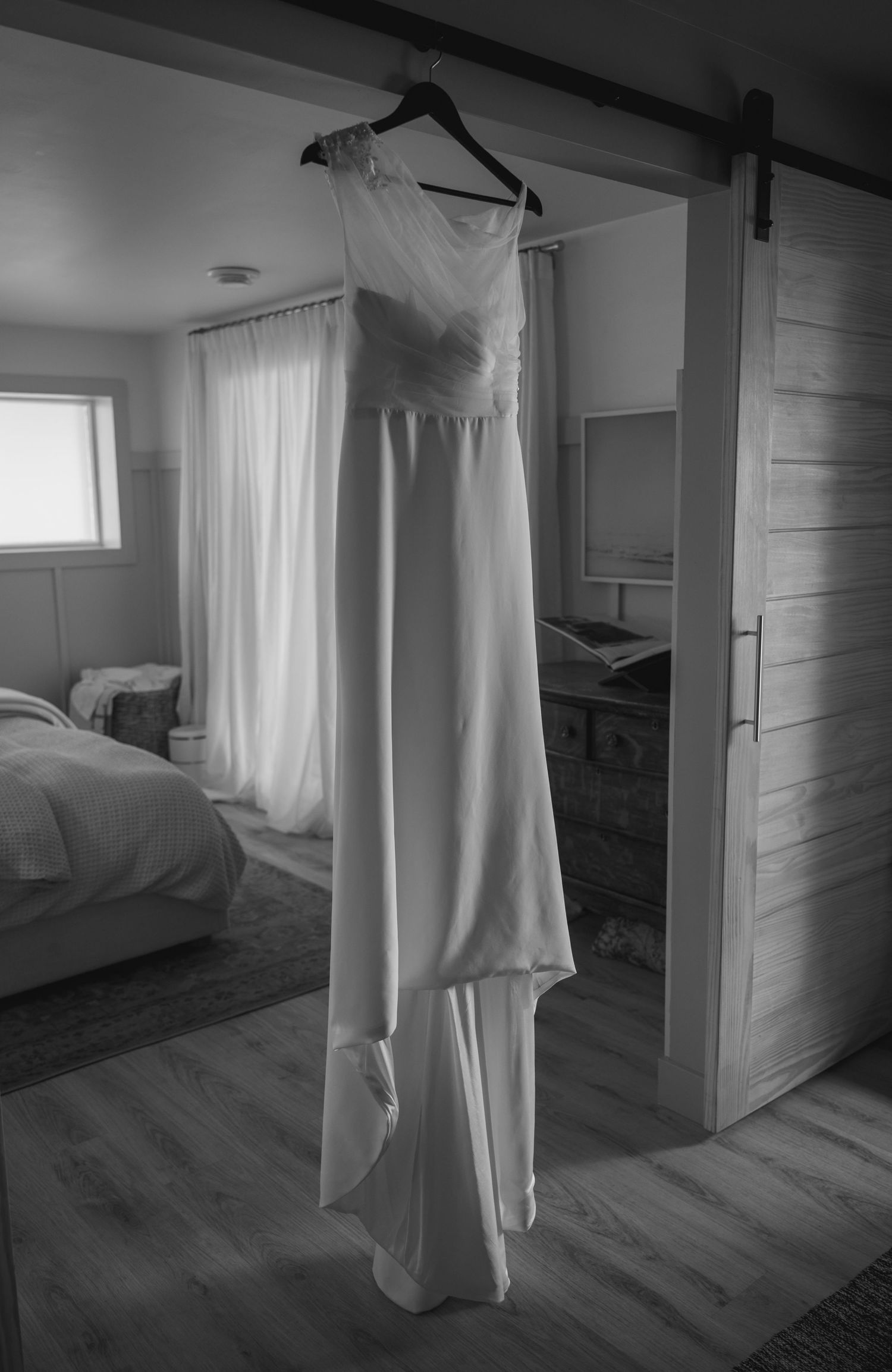 A white wedding dress hangs ethereally in a sunlit room with hardwood floors.