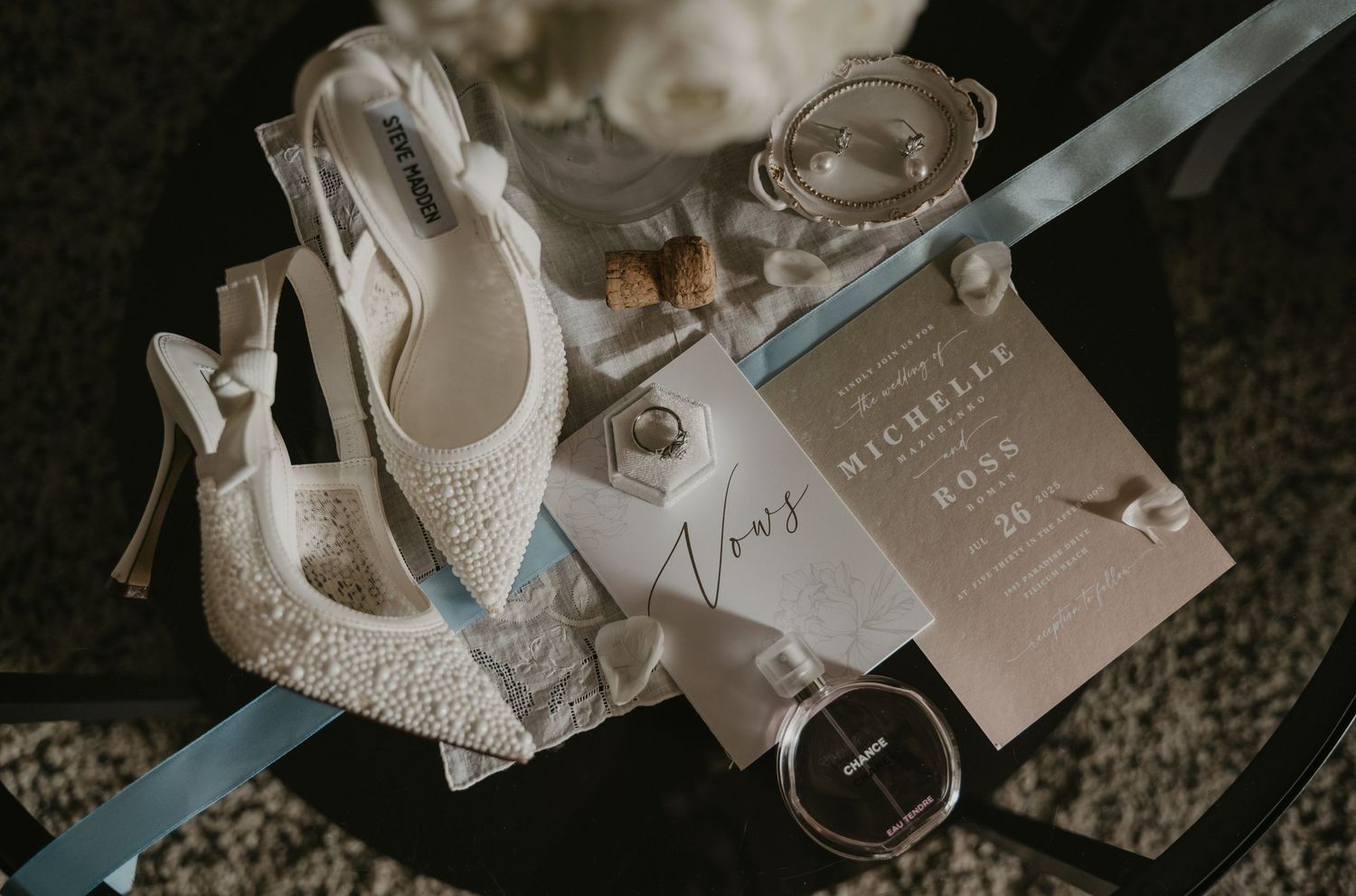 Elegant white lace bridal heels and wedding invitation cards arranged on dark textured surface in soft lighting.