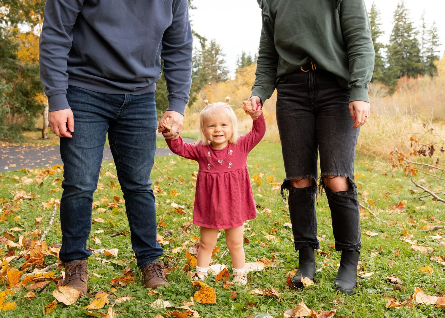 A small child in a red dress stands joyfully between two people on grass covered with autumn leaves.