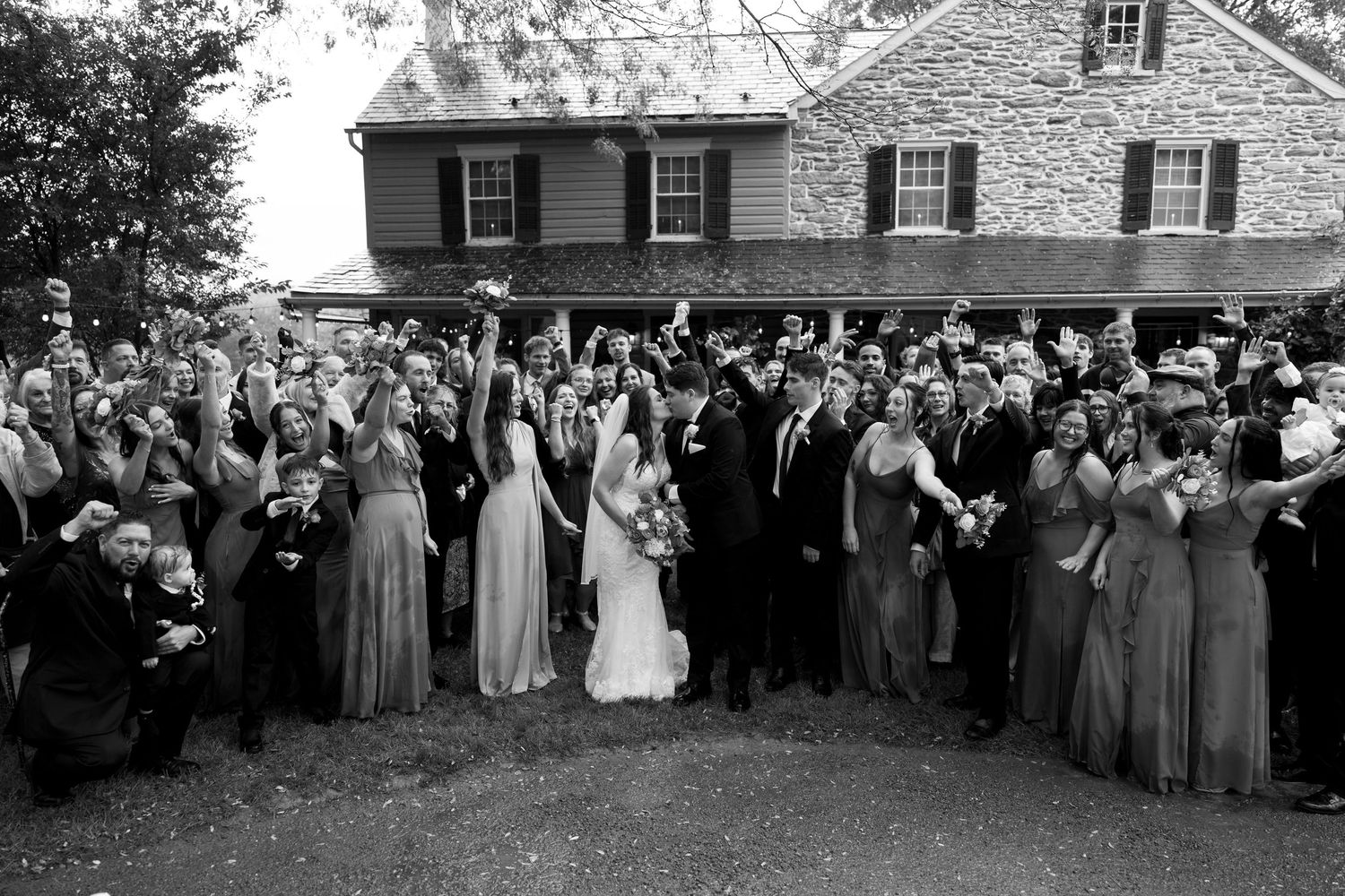 Wedding party and guests pose together in front of a stone house wearing sage green bridesmaid dresses.