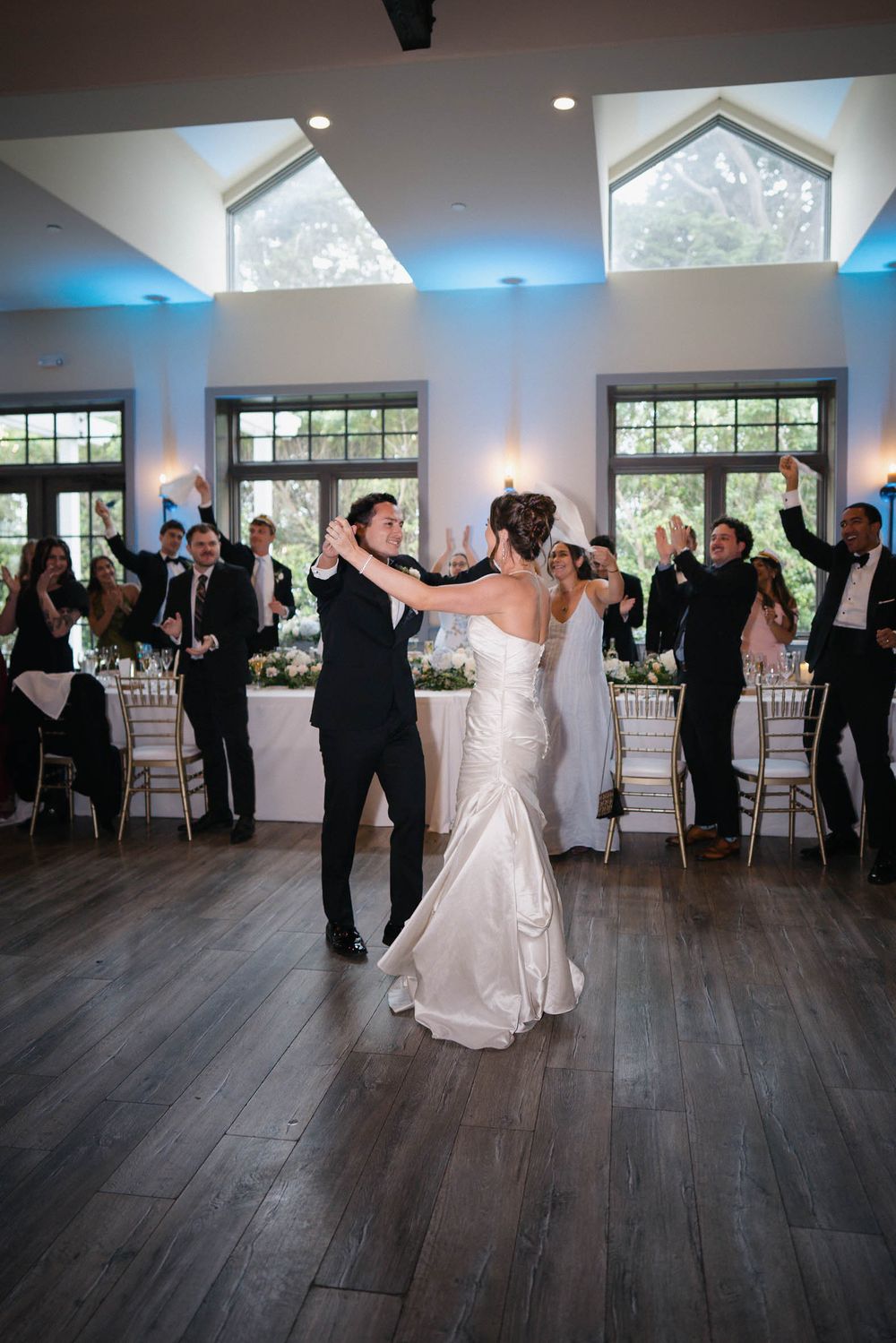 Wedding guests dance and celebrate on a hardwood floor in an elegant reception venue with large windows.