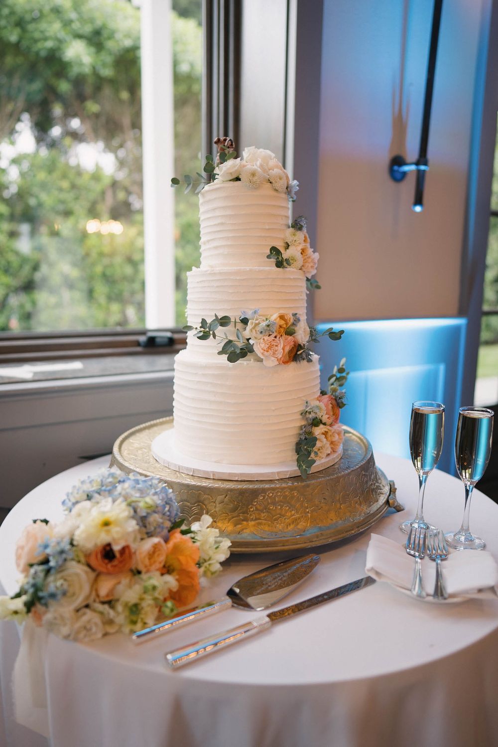 White tiered wedding cake decorated with fresh flowers displayed on a table near windows.