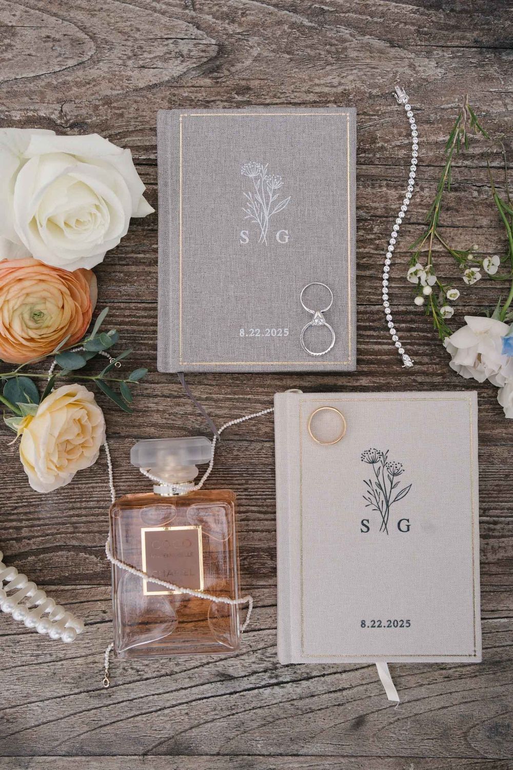 Wedding invitation books with floral design displayed with roses, perfume bottle, and pearl necklace on rustic wooden surface.