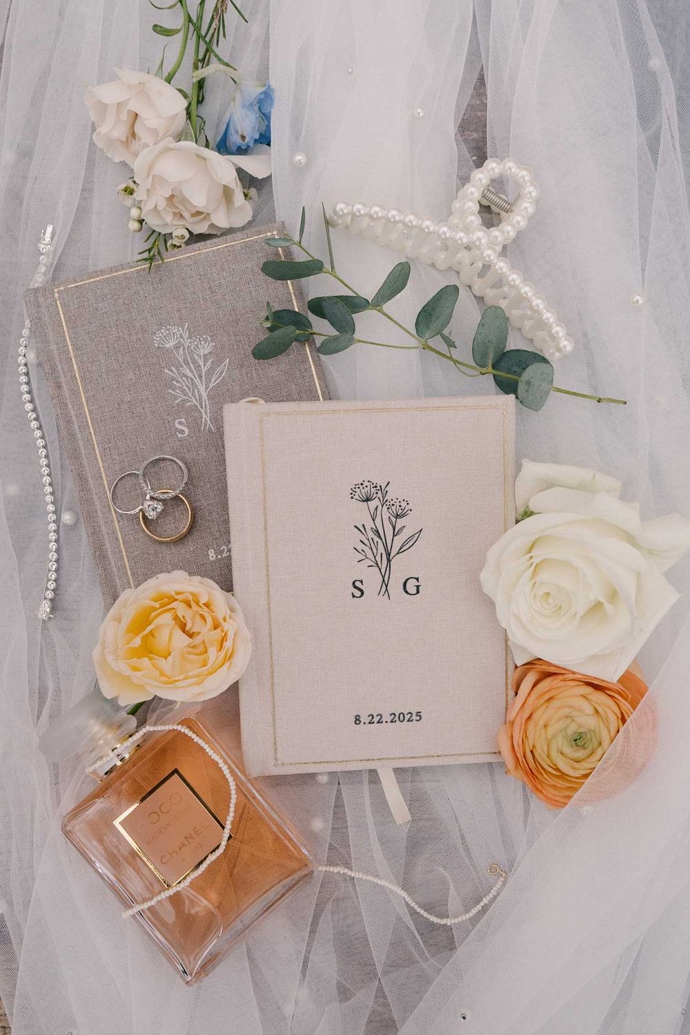 Wedding details flatlay with perfume bottle, rings, invitation, and white roses on delicate tulle fabric background.